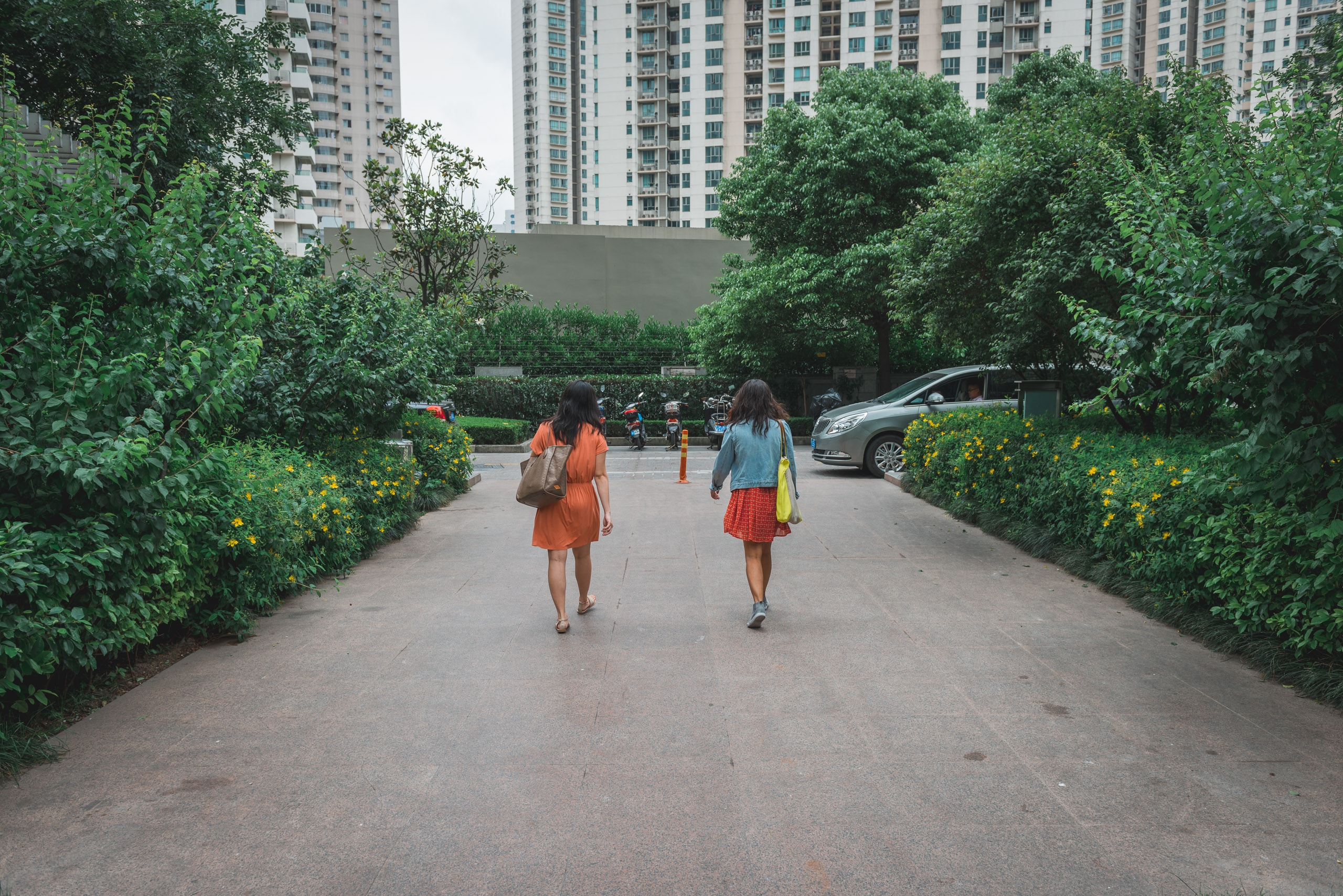 Two women walk down a tree-lined pathway between lush green hedges in an urban residential complex, with apartment towers visible in the background under overcast sky
