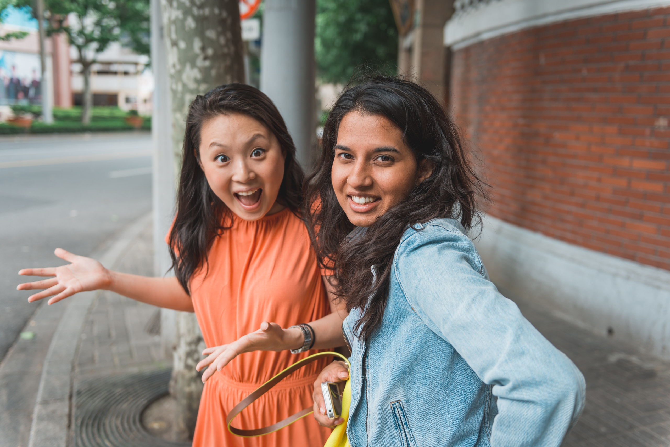 Two young women pose cheerfully on a urban sidewalk, one wearing an orange dress with arm outstretched and the other in a denim jacket, with brick buildings and metal pillars behind them