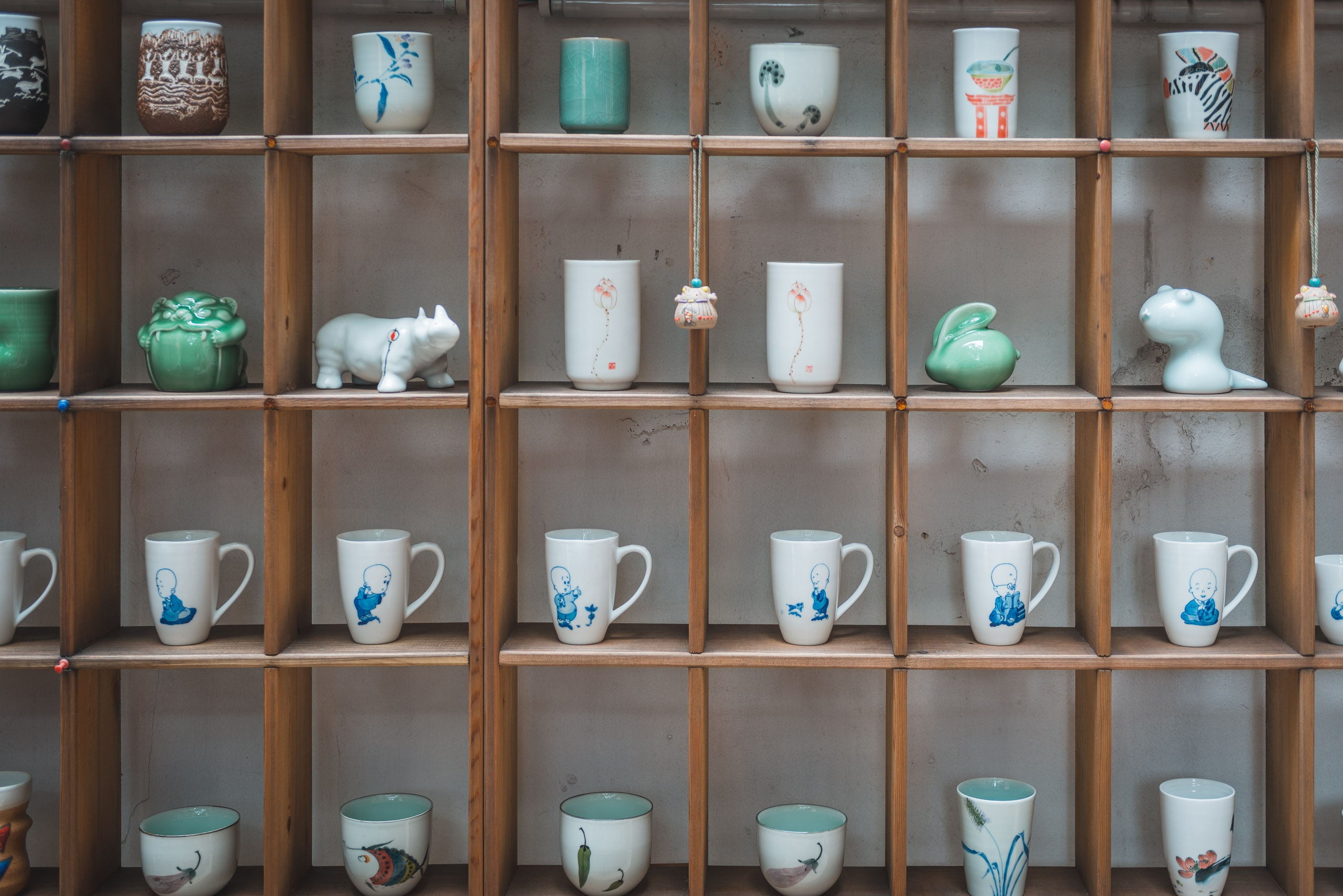 Wooden shelving unit displays an organized collection of ceramic vessels including painted porcelain mugs, decorative bowls, and artistic vases with blue and green accents