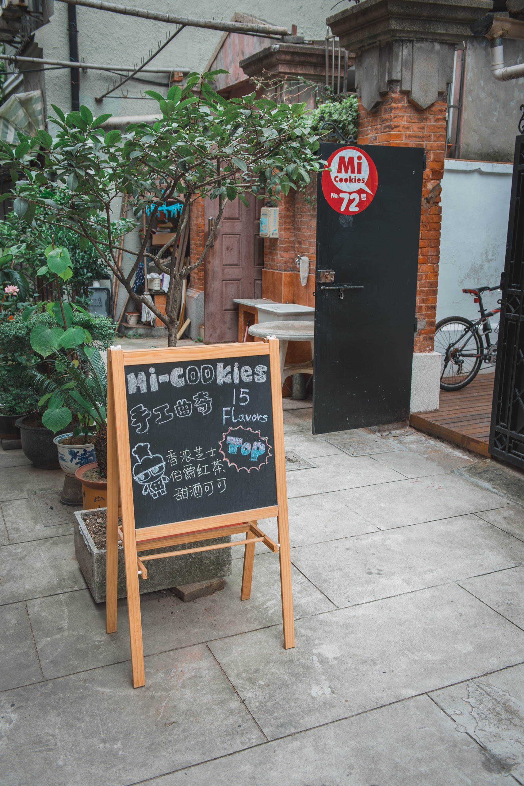 A wooden A-frame chalkboard advertises Mi-cookies outside a brick storefront with a black door, potted plants, and a bicycle visible in the narrow alleyway
