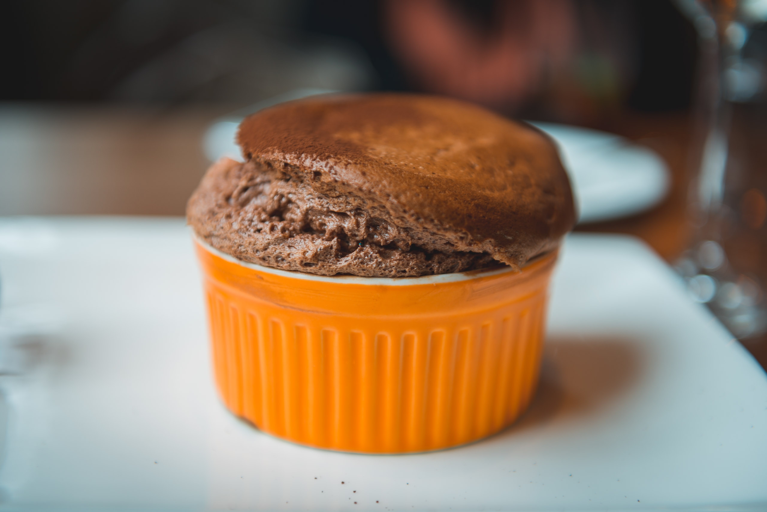 A risen chocolate soufflé in a bright orange ramekin sits on a white plate with soft natural lighting casting gentle shadows
