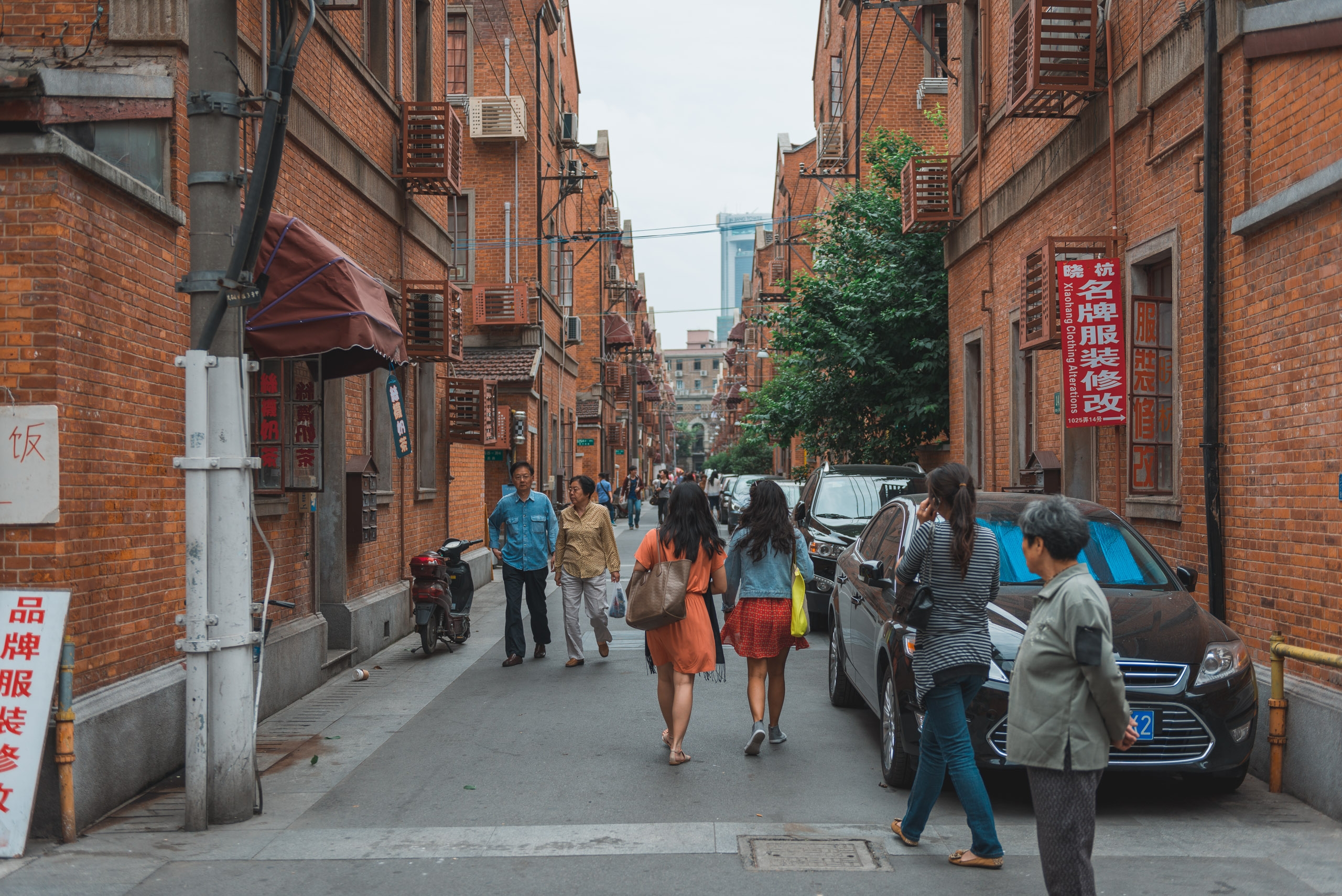 A narrow brick alley in Shanghai lined with residential buildings features pedestrians and parked cars beneath overcast skies, with red Chinese signage visible on the weathered walls