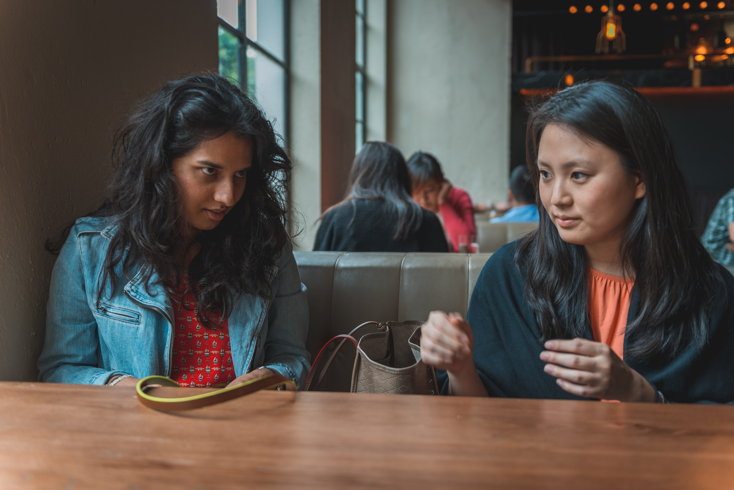 Two women sit at a wooden table in an indoor venue, one in a denim jacket and the other in an orange top, engaged in conversation while other patrons blur in the background