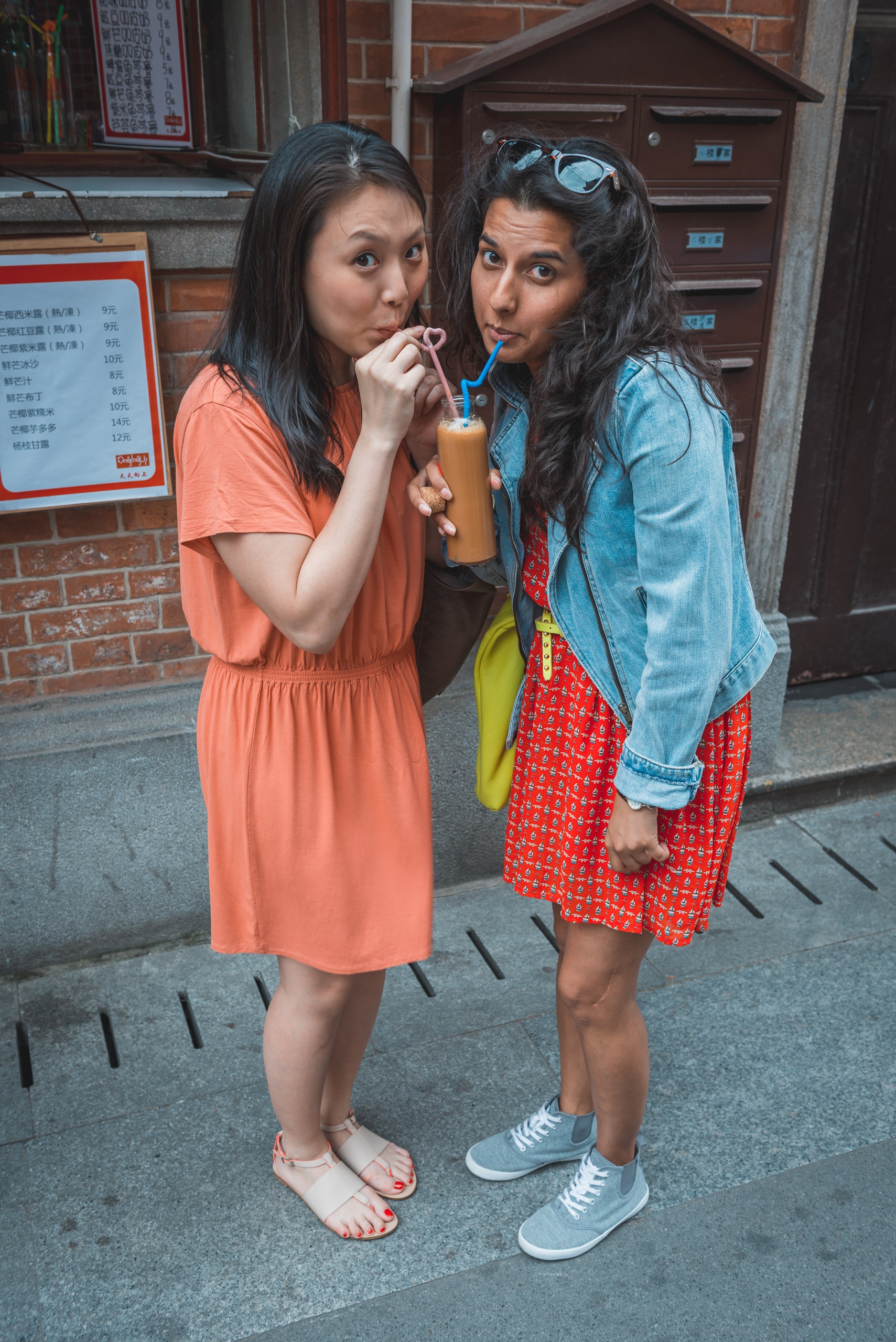 Two young women stand on a narrow street outside a traditional brick building, sharing a beverage through a single straw while wearing casual, colorful clothing