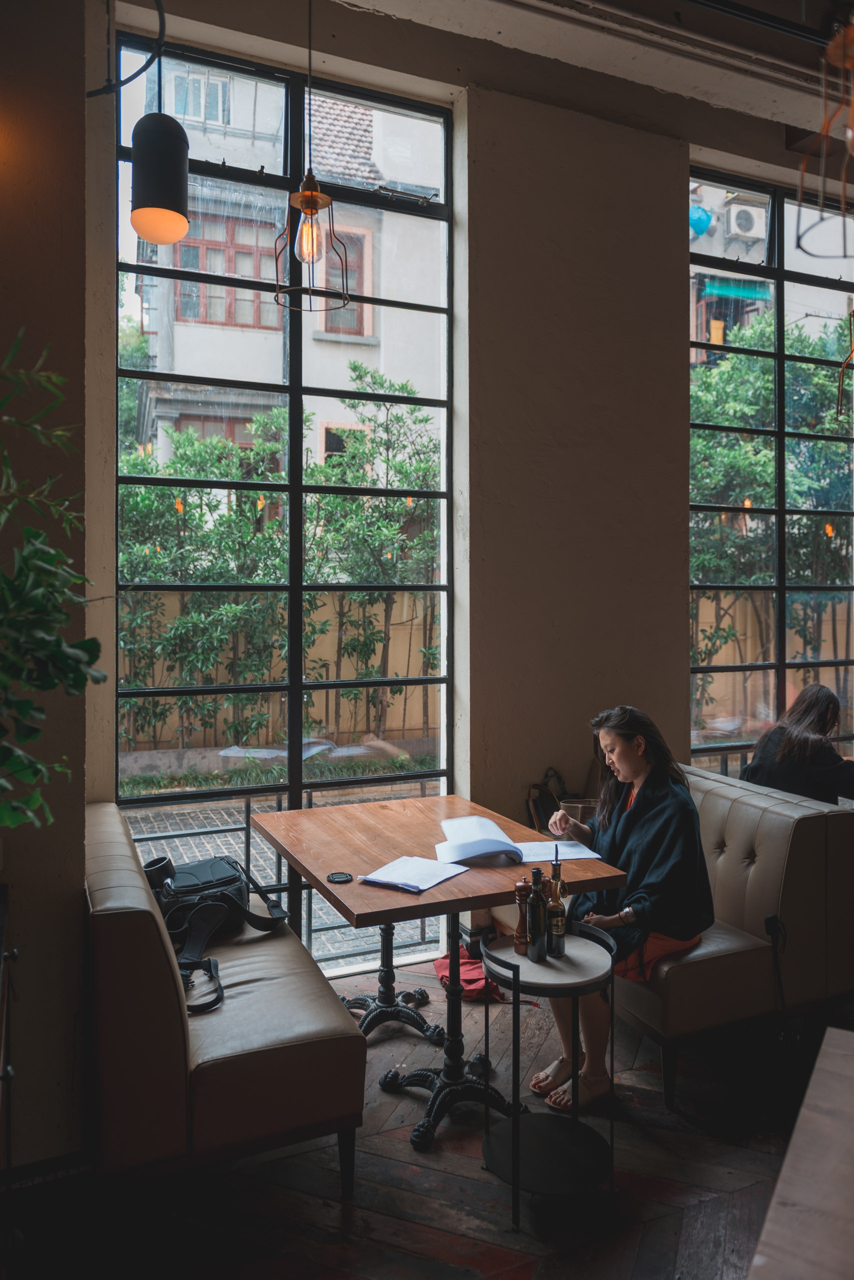 A woman sits at a wooden desk working in a modern loft space with large industrial windows overlooking a green urban courtyard and neighboring buildings.