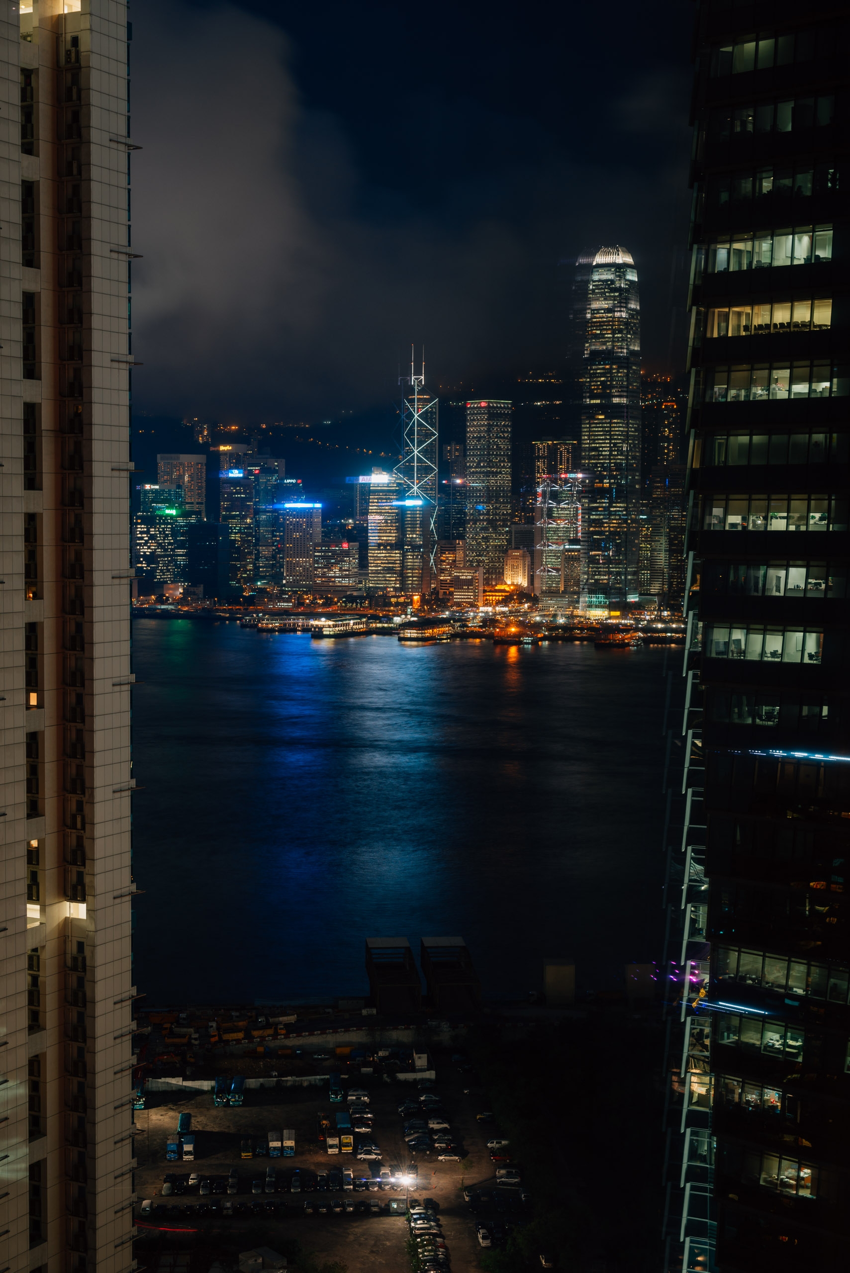 Hong Kong's illuminated skyline reflects across Victoria Harbour at night, framed by residential and office buildings in the foreground with lit windows