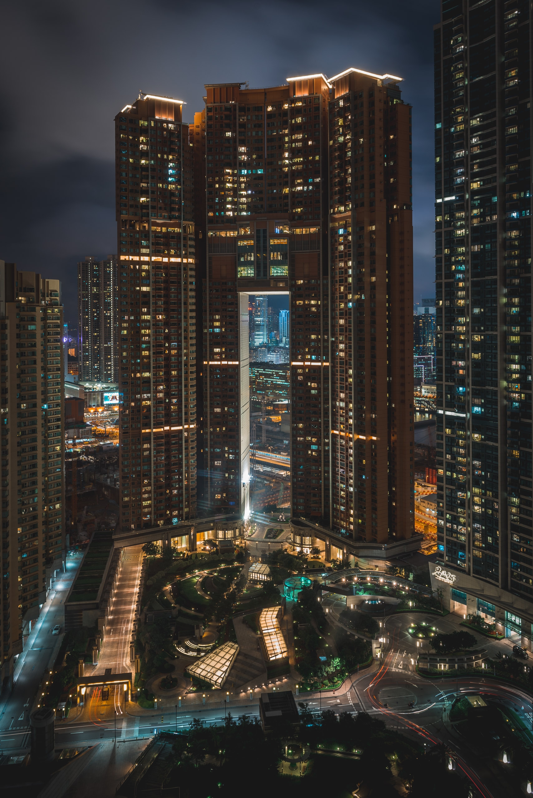 Illuminated high-rise towers frame a lit courtyard plaza at twilight, with warm interior lights glowing against the darkening blue sky above the urban Hong Kong skyline