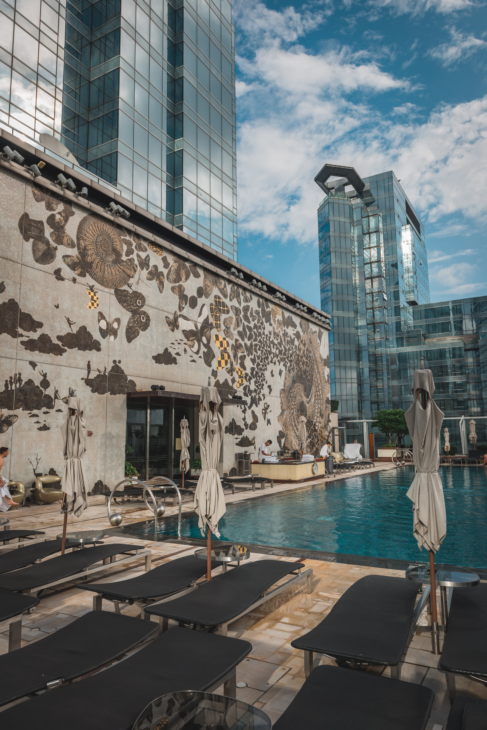 A modern rooftop pool area features black loungers and umbrellas beside a turquoise pool, with an ornate mural-covered heritage building and contemporary glass skyscrapers under a bright blue sky