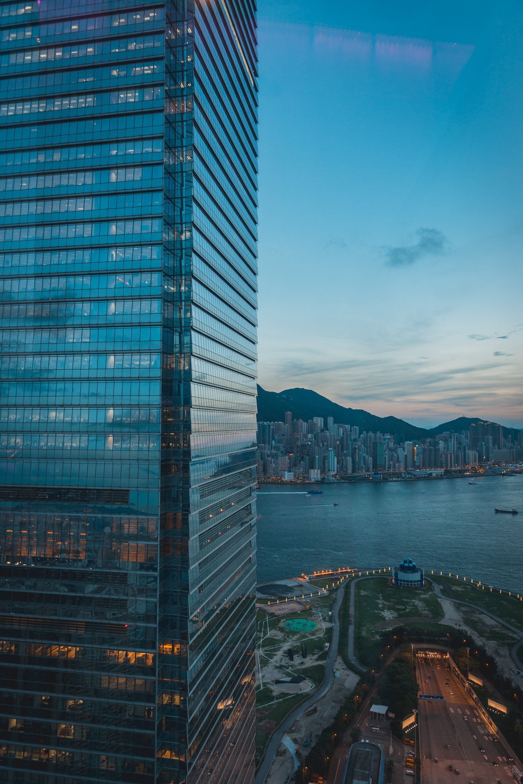 Modern glass tower rises beside a harbor at dusk, with illuminated waterfront buildings and mountains visible across Victoria Harbour under a twilight sky