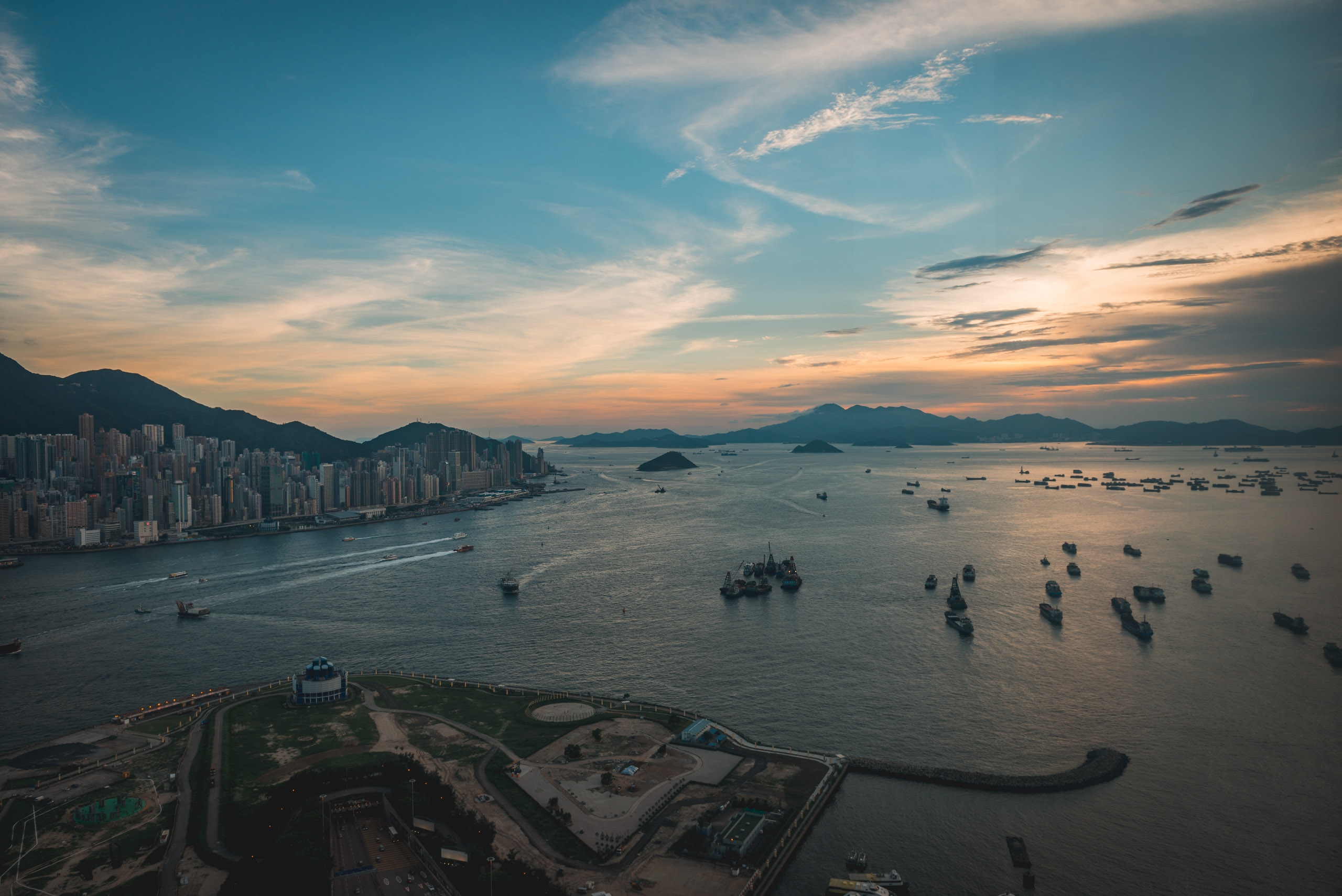 Dense urban skyline of Hong Kong rises beside a harbor filled with anchored fishing vessels and cargo ships, framed by distant mountains under a golden sunset sky