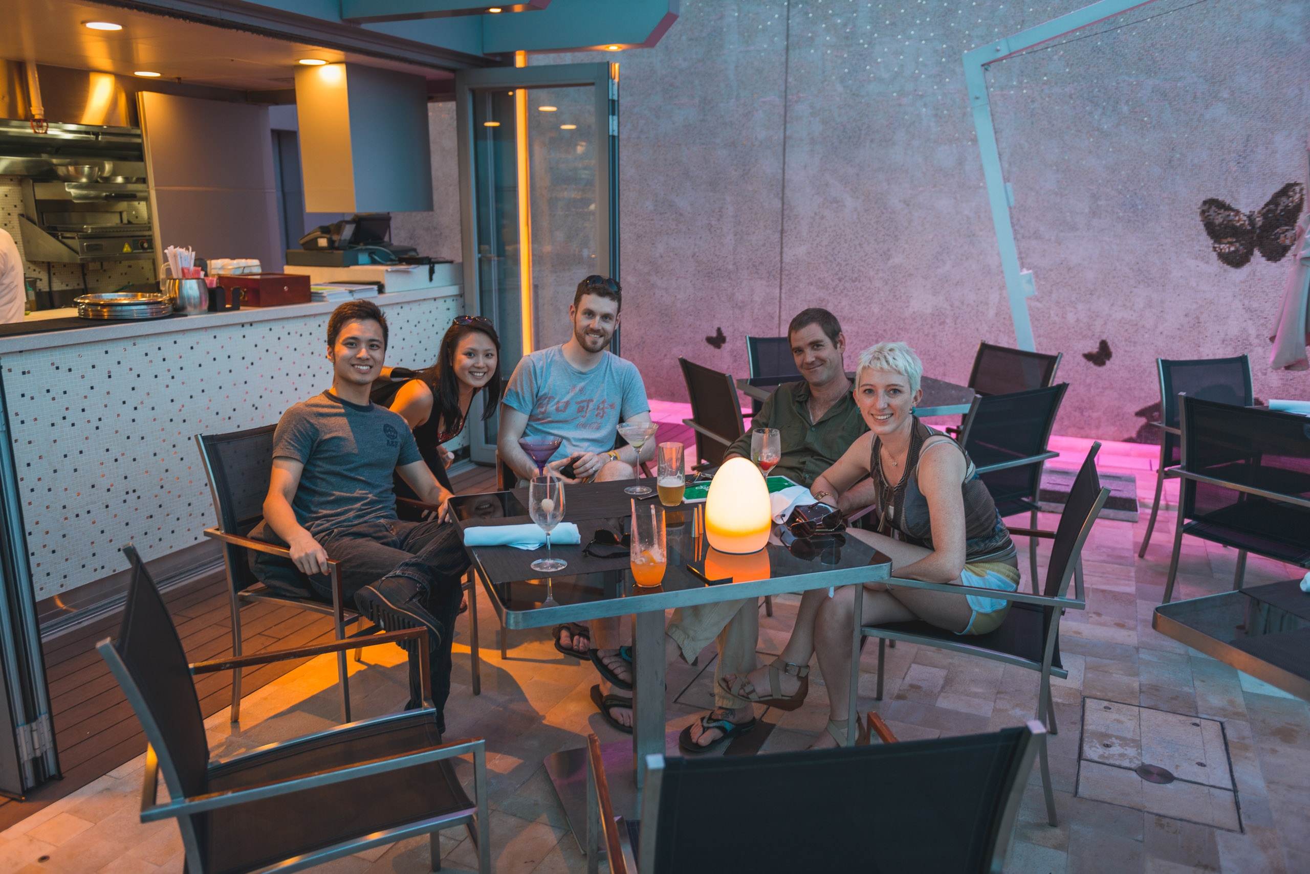 Five people gather around a low table in a modern lounge space, illuminated by warm orange and pink neon lighting, with laptops and a glowing lamp visible among casual seating