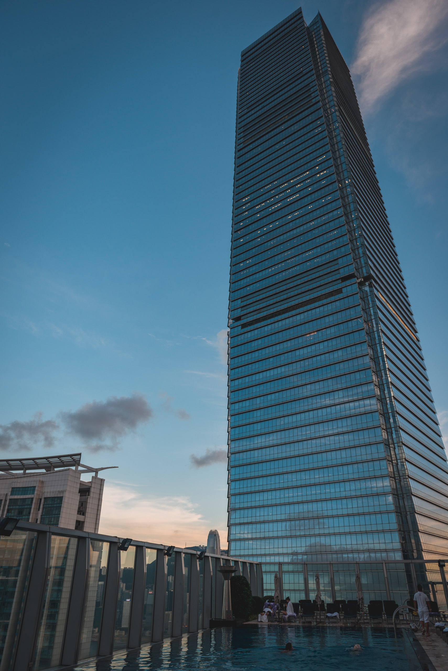 A tall glass-fronted residential tower rises against a clear blue sky, with a rooftop swimming pool and adjacent modern building visible in the foreground below