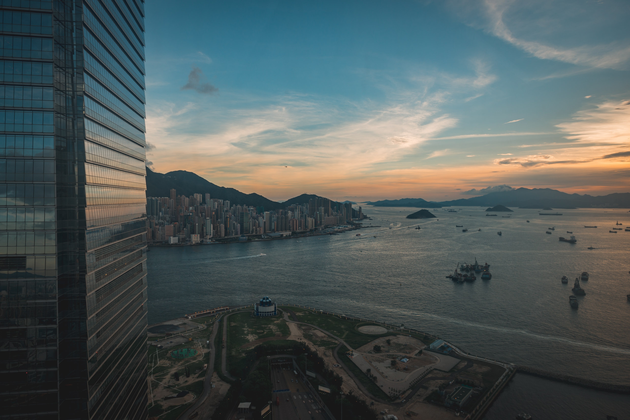 Modern glass skyscrapers frame a sunset view over Victoria Harbour, with dense waterfront buildings, anchored vessels, and distant mountains bathed in warm golden light