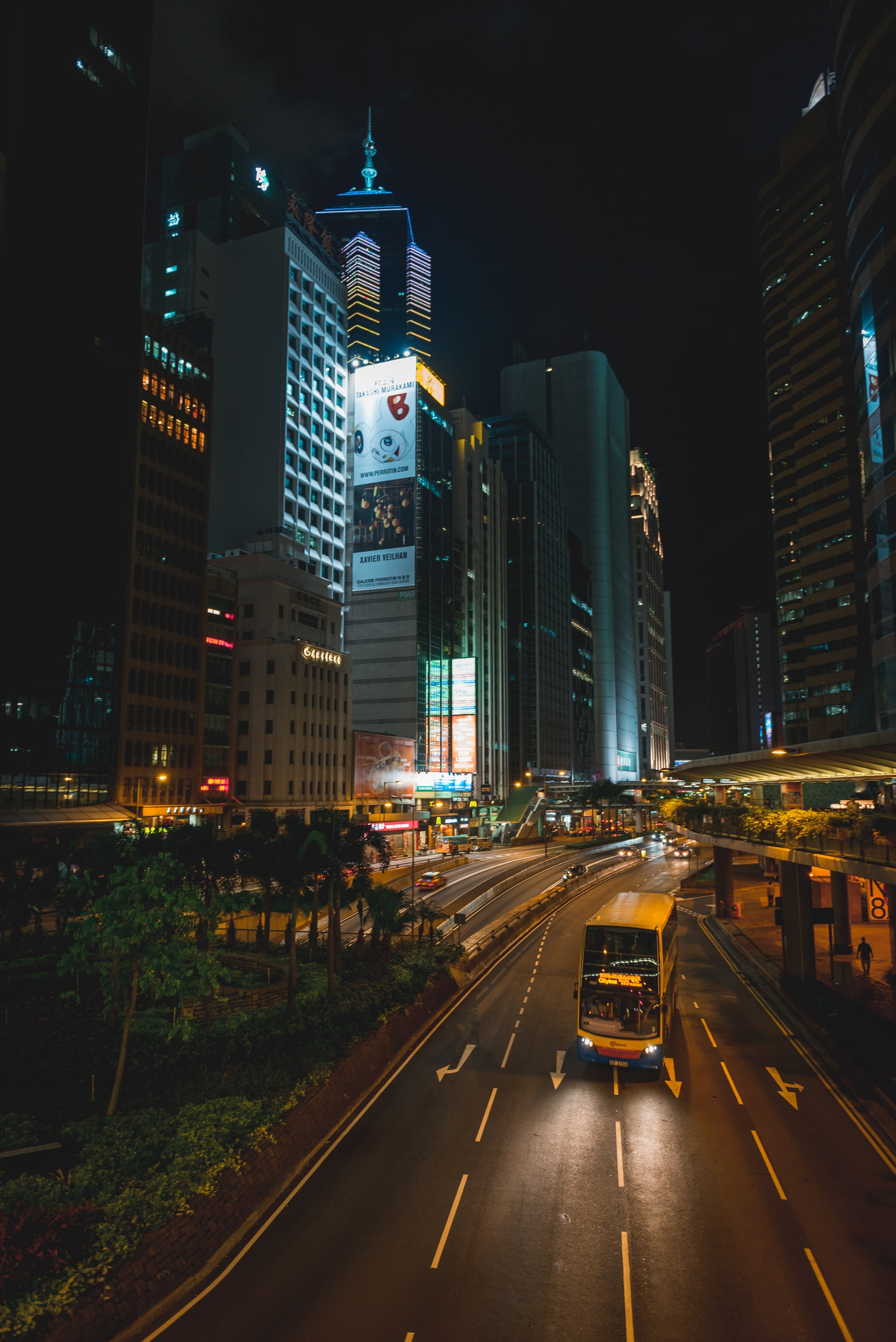 Yellow taxi drives down a wet Hong Kong street at night, flanked by illuminated skyscrapers and glowing advertisements under dark skies