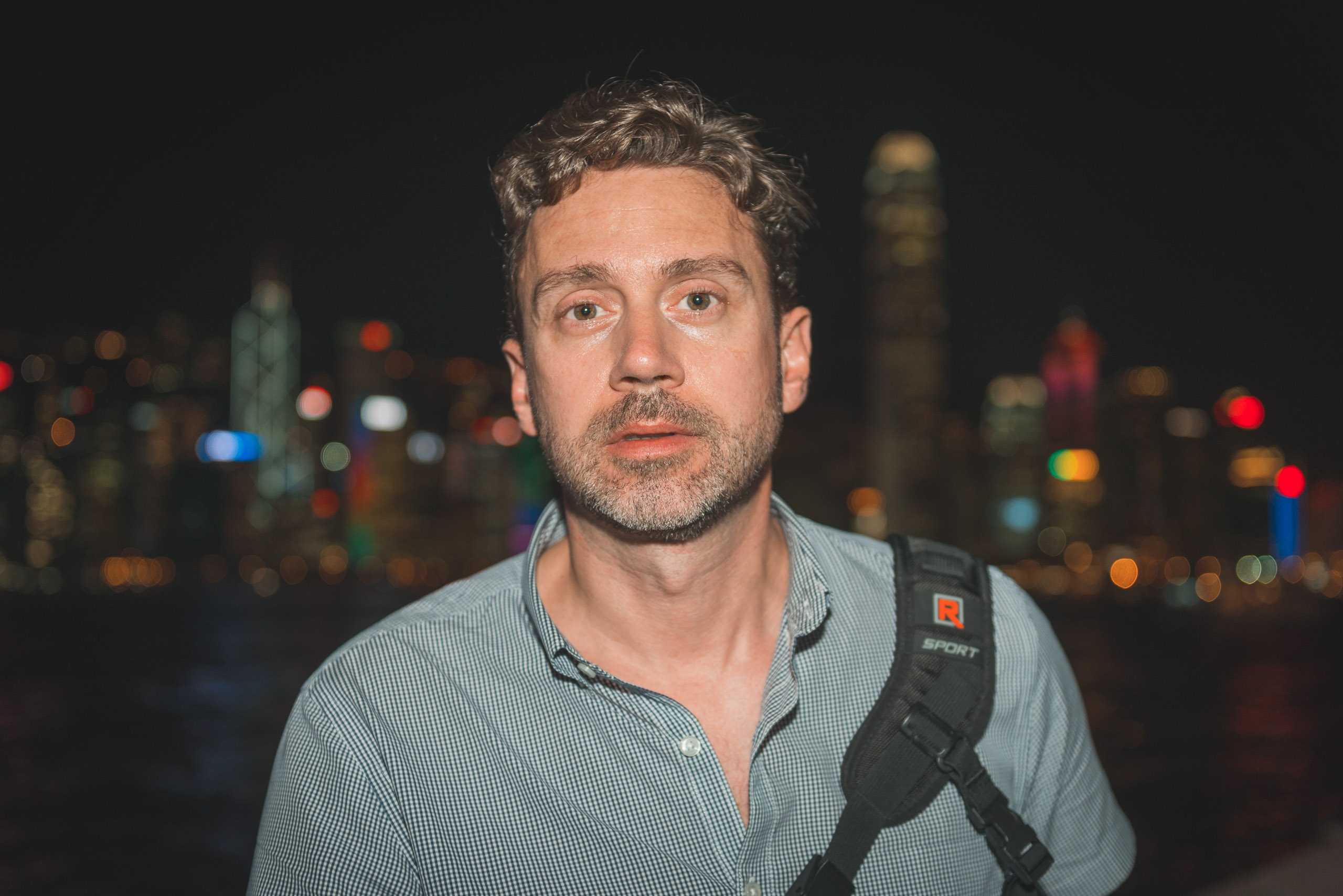 Man wearing a light blue shirt and camera strap poses against a blurred nighttime cityscape with colorful bokeh lights from distant buildings