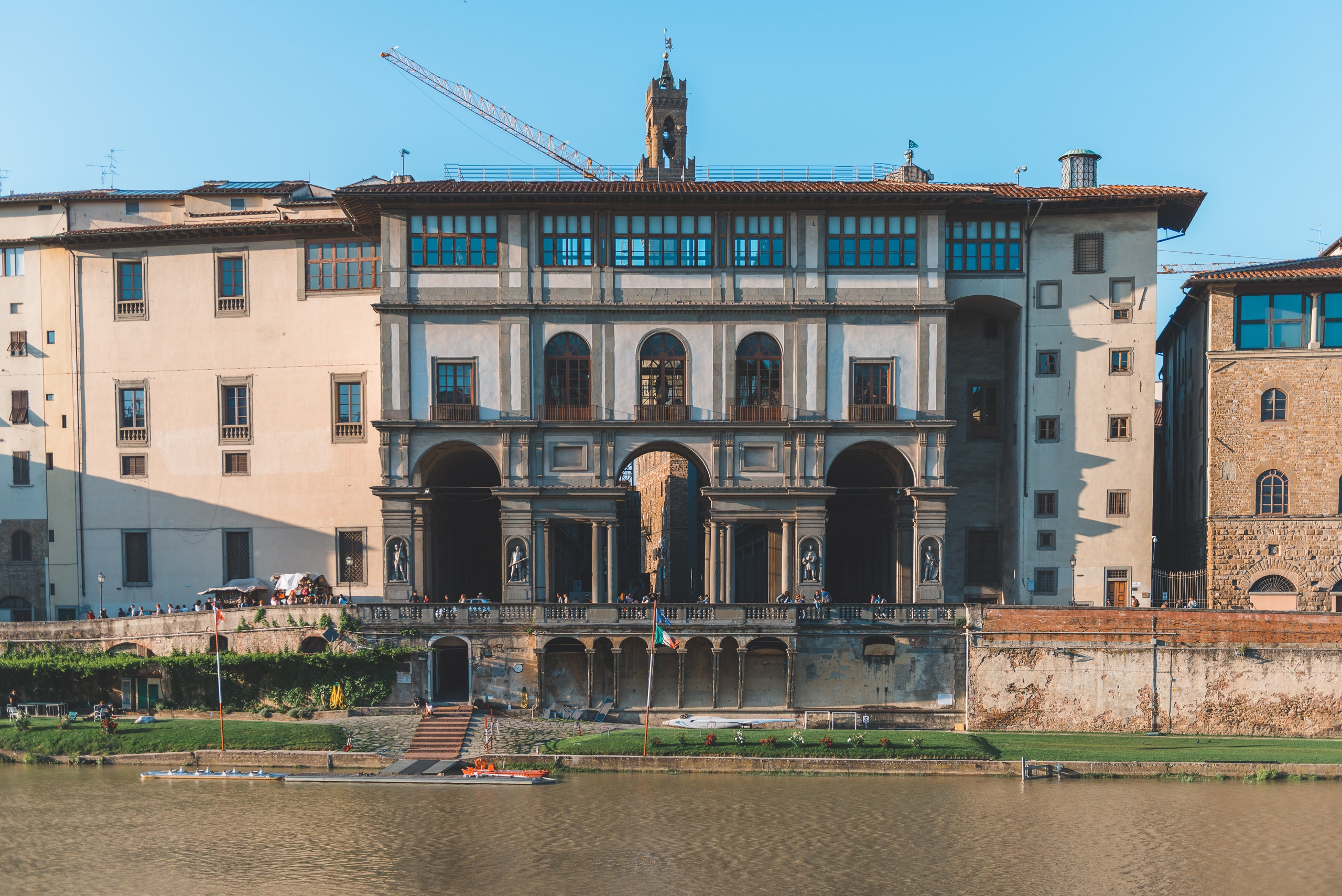 A Renaissance palazzo with symmetrical arched openings faces the Arno River in Florence, its cream and gray stone facade gleaming under clear sunlight with surrounding period architecture visible