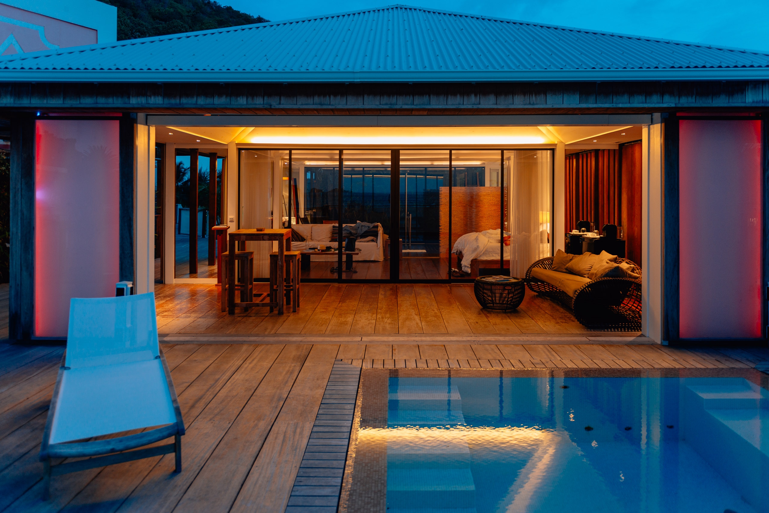 A modern resort suite glows with warm interior lighting at dusk, featuring wooden decking, floor-to-ceiling windows, and a illuminated swimming pool reflecting the blue twilight sky