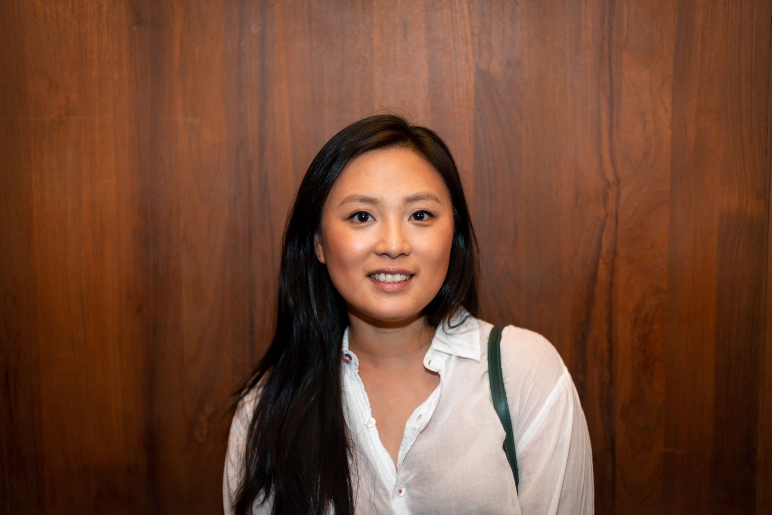 A woman with long black hair wearing a white collared shirt smiles at the camera against a rich brown wood-paneled background with warm, even lighting