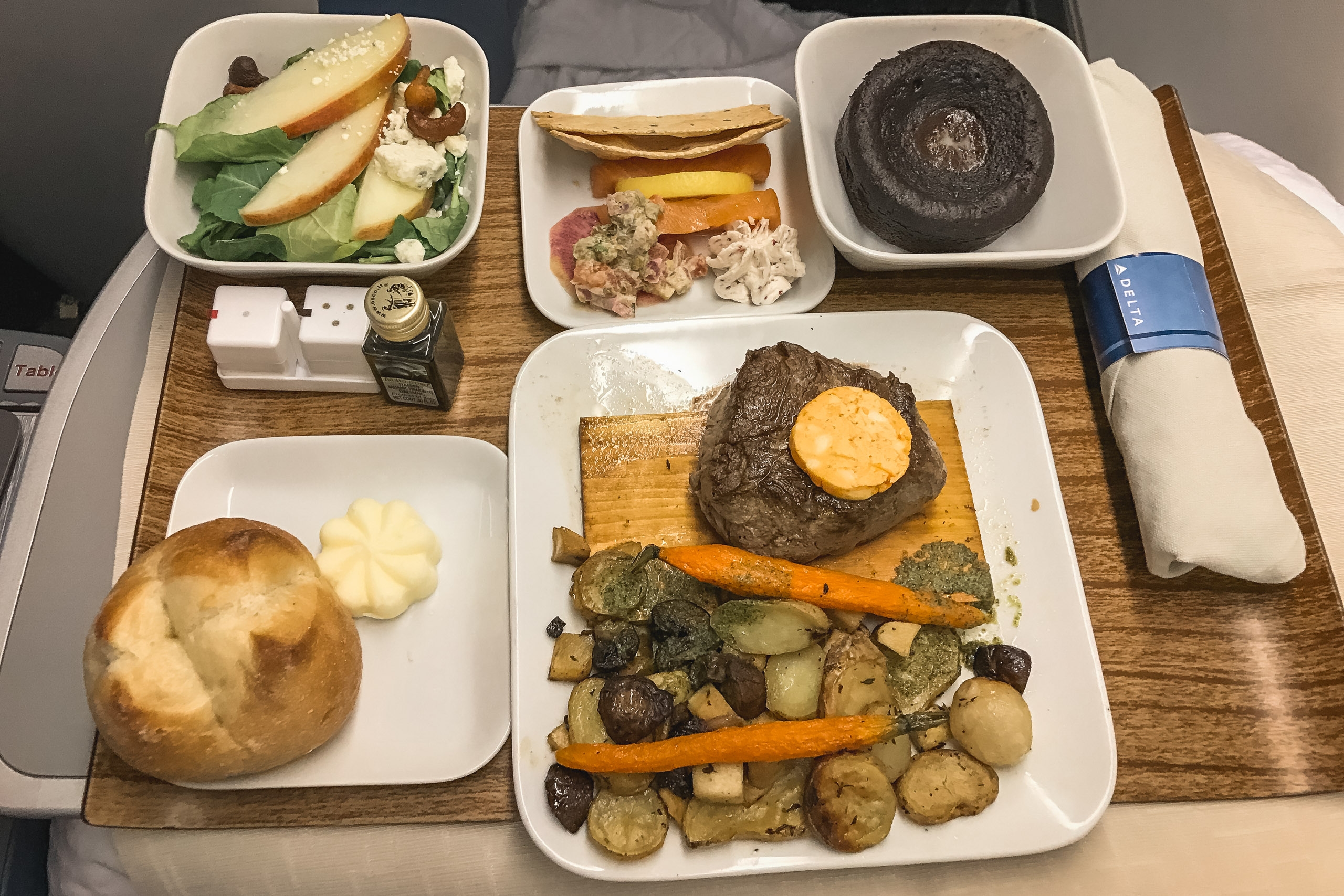 Airline meal tray features a grilled steak with butter and roasted vegetables, fresh bread roll, green salad with apple slices, and dark sauce, arranged on white plates atop a wooden serving board