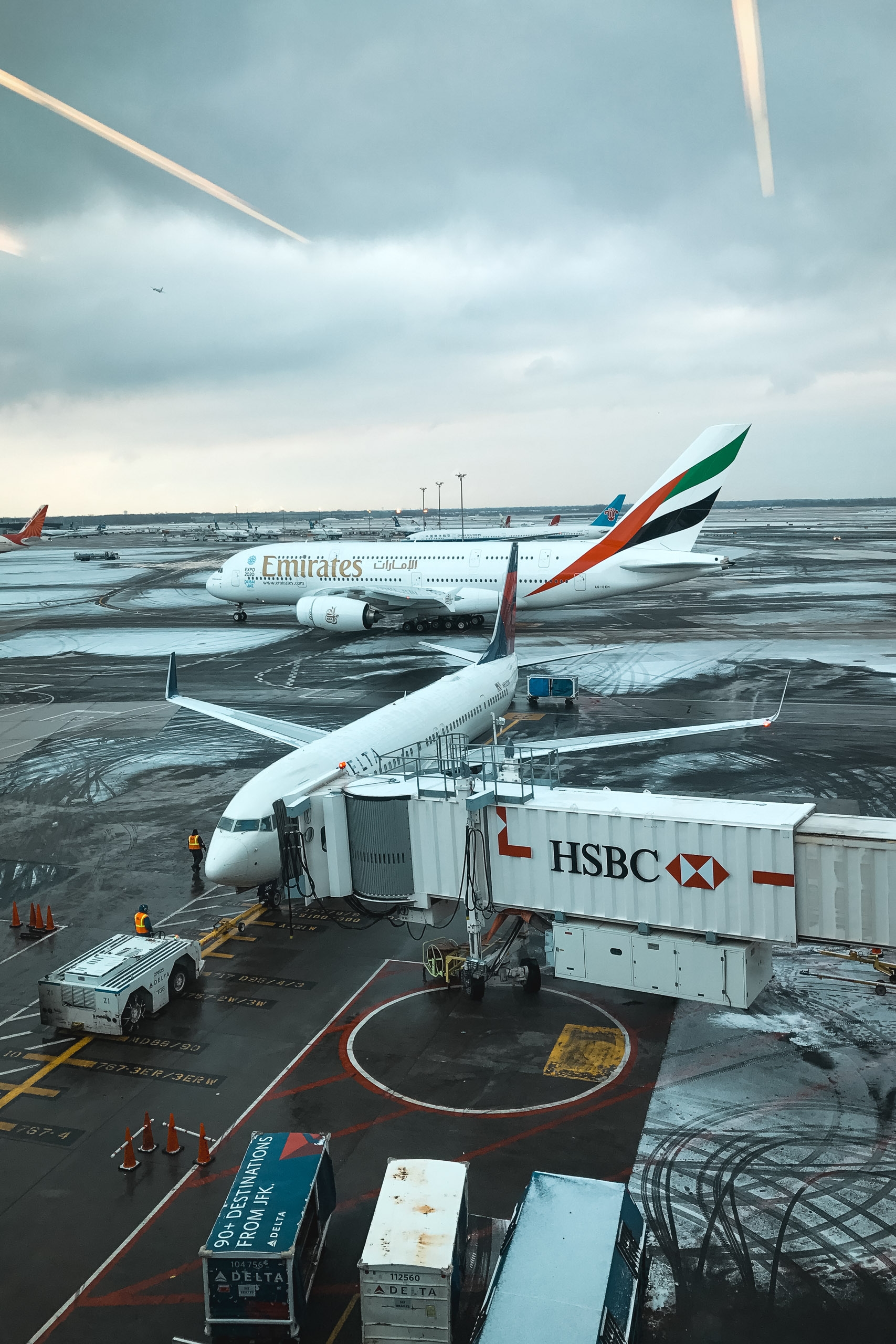 An Emirates aircraft docked at an airport gate with an HSBC-branded jet bridge and ground service vehicles clustered beneath its fuselage under overcast skies
