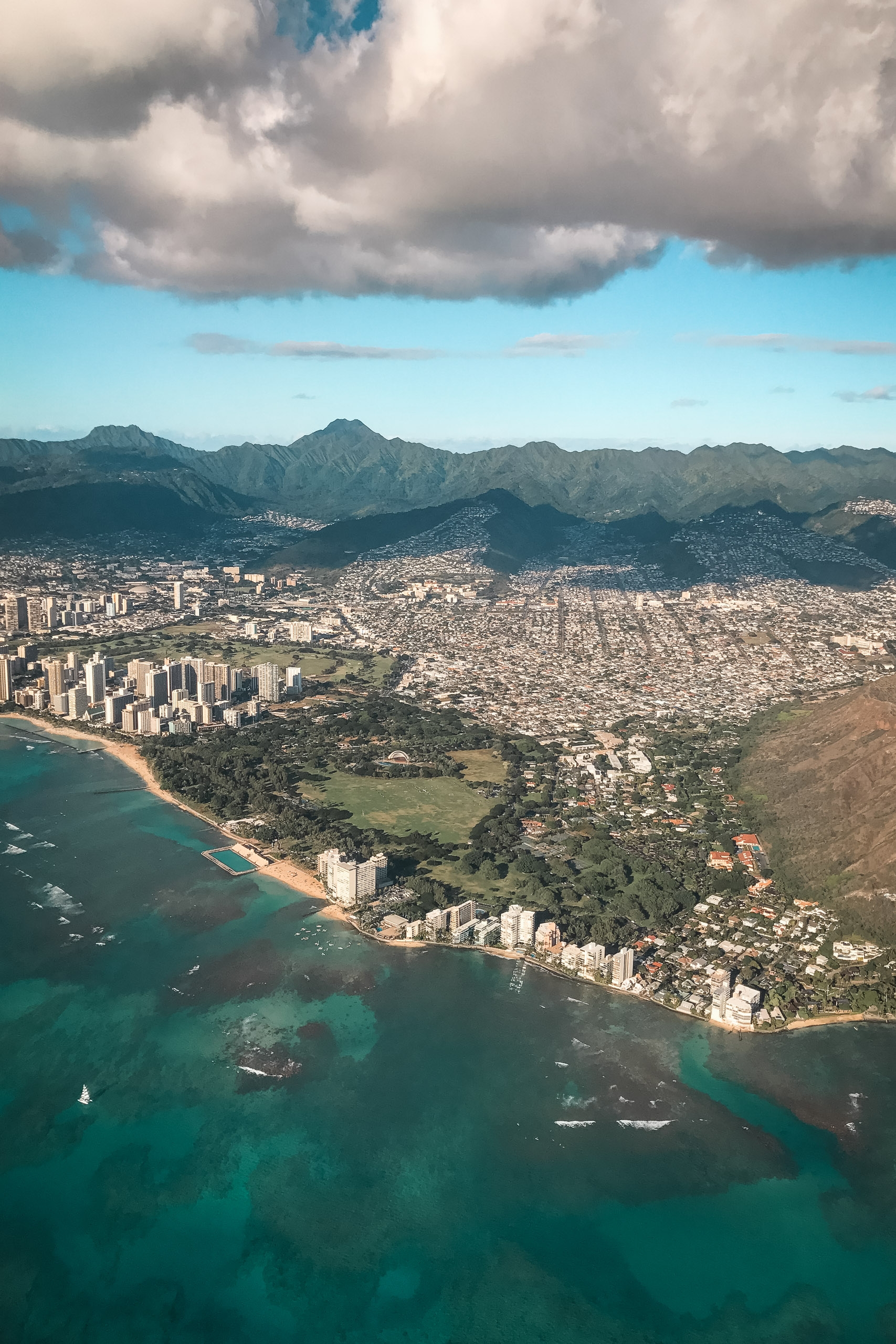 Aerial view of Oahu's coastal landscape featuring turquoise waters, a curved sandy beach lined with high-rise buildings, lush green valleys, and volcanic mountains beneath dramatic white clouds