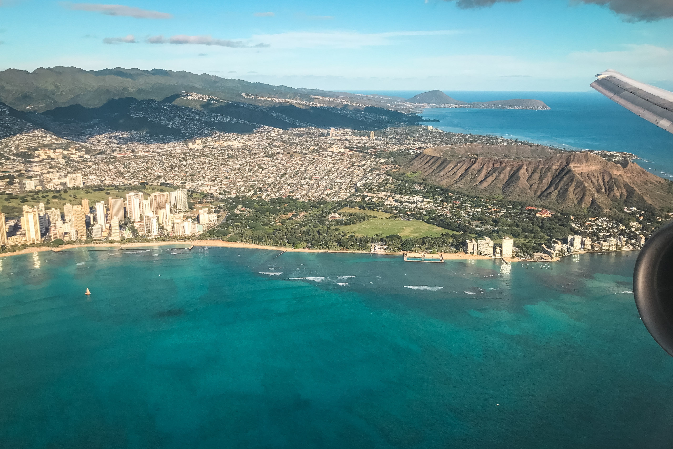 Aerial view of Waikiki Beach and Diamond Head crater from an aircraft, showing turquoise ocean waters, high-rise hotels, green valleys, and distant volcanic mountains under bright daylight
