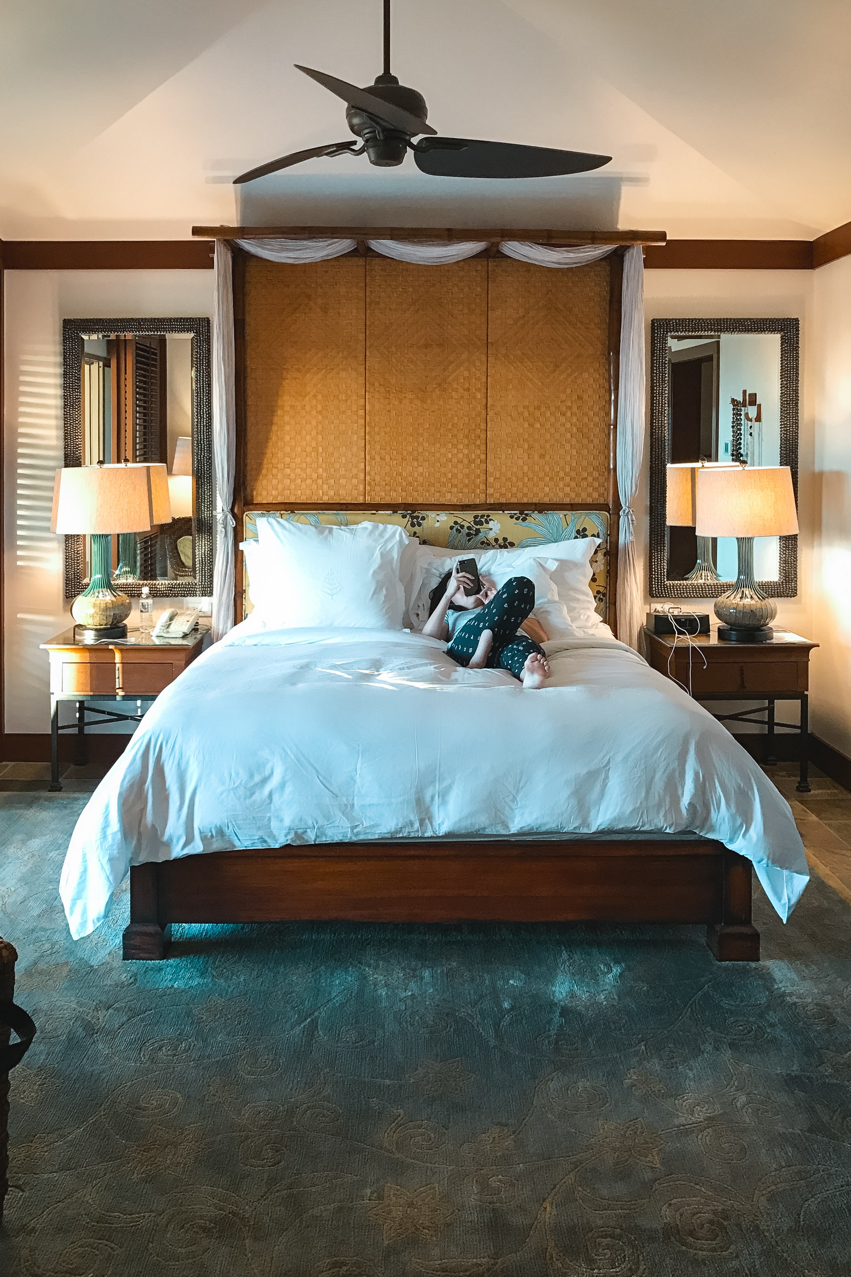 A luxurious four-poster bed with crisp light blue bedding sits centered in an elegant bedroom, flanked by matching wooden nightstands with brass lamps beneath a ceiling fan