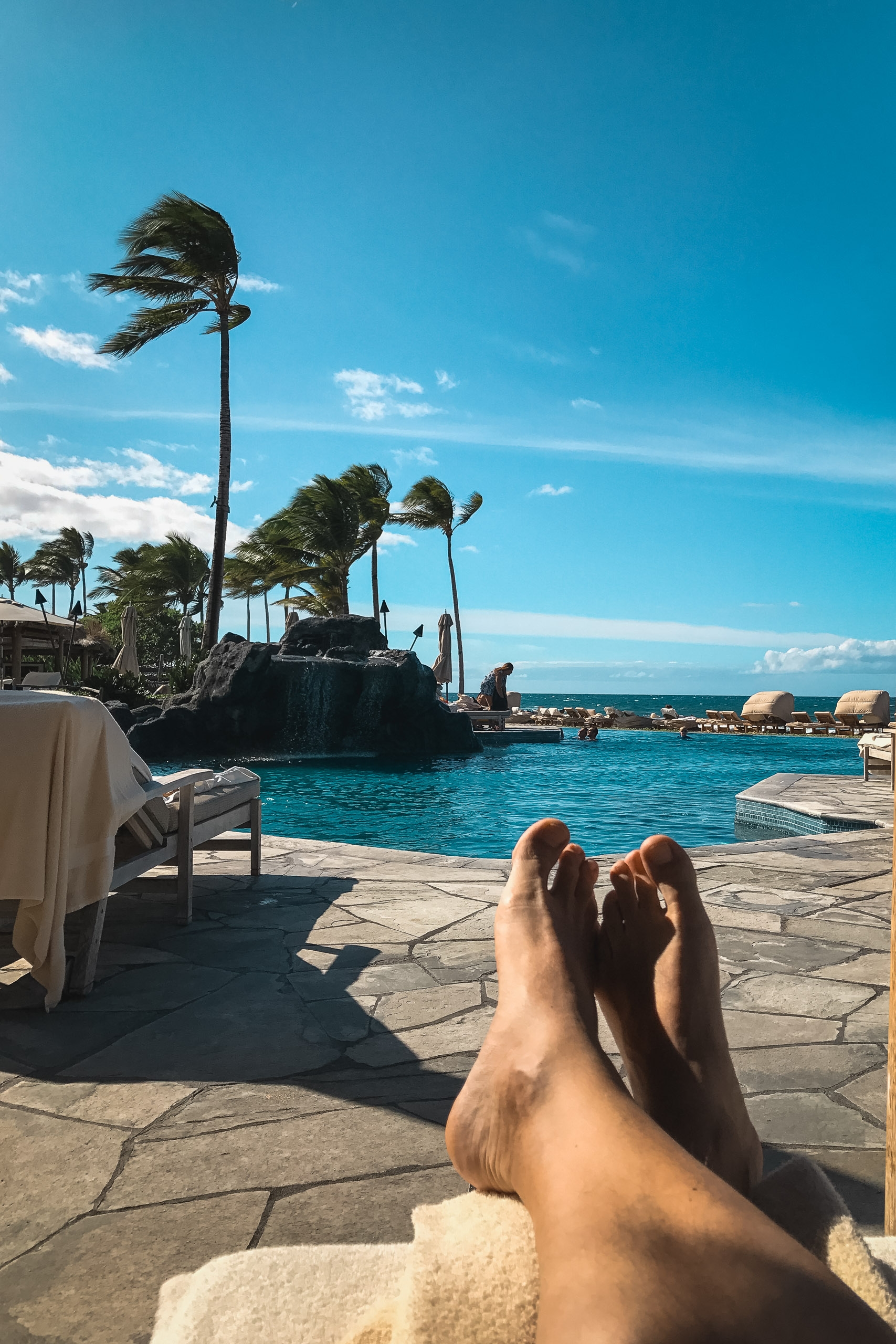 A person's bare feet rest on a sunlit stone pool deck overlooking a turquoise lagoon with swaying palm trees and white umbrellas in the background under a clear blue sky