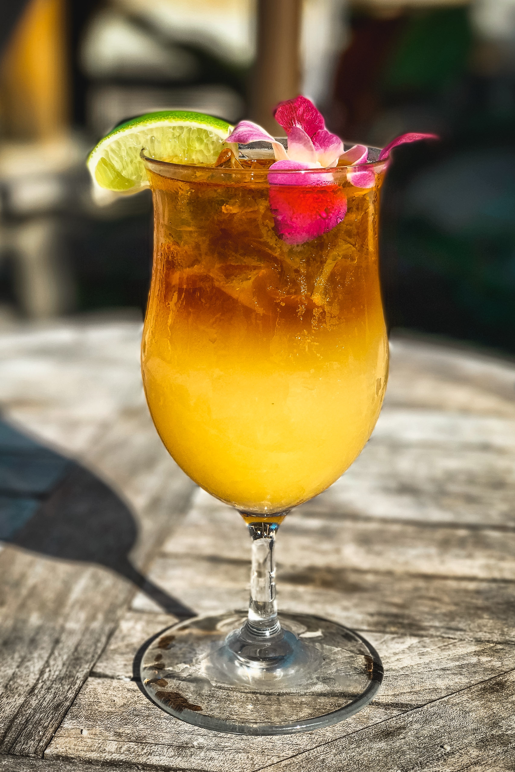 A golden tropical cocktail garnished with a lime wheel and vibrant pink flower sits on a weathered wooden surface in bright sunlight