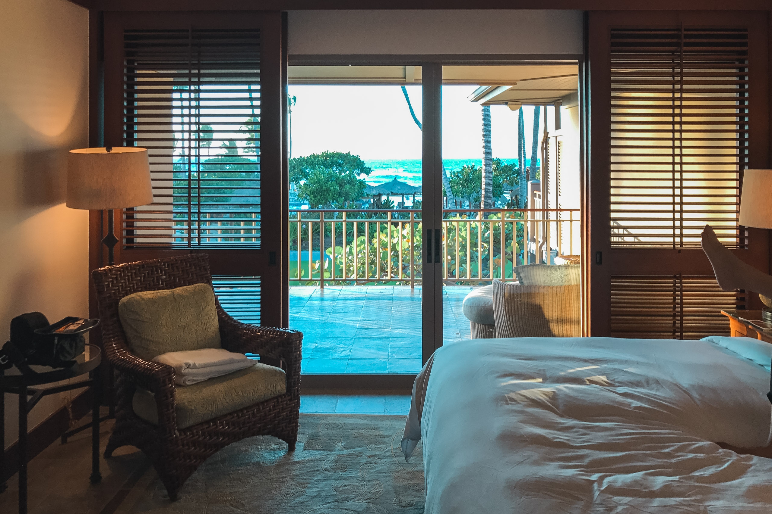 Luxury hotel bedroom overlooks turquoise ocean and tropical landscape through open glass doors, with wicker chair and warm lamp lighting the interior space