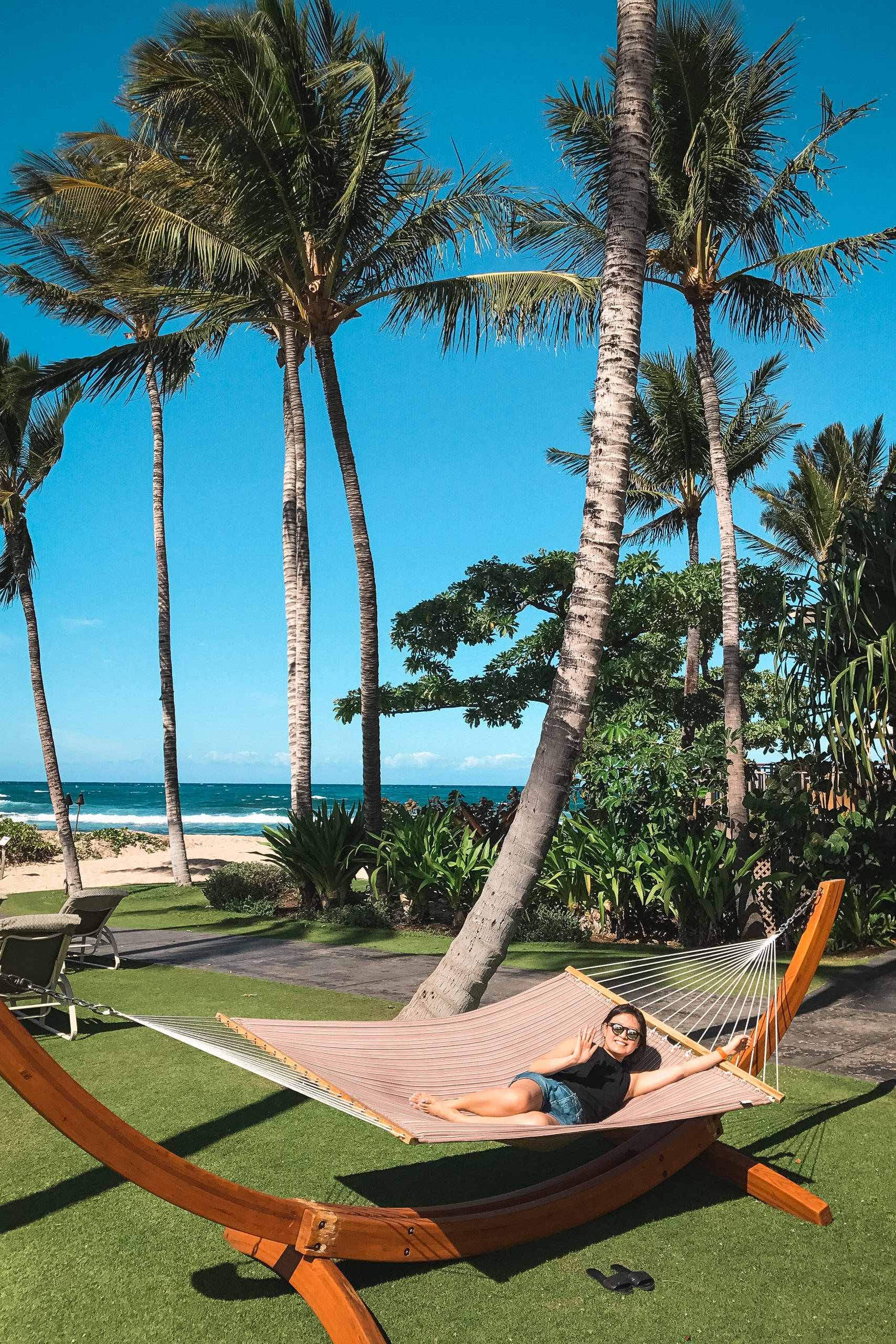 A woman relaxes in a hammock on a manicured beachfront lawn, surrounded by tall palm trees with the ocean and clear blue sky visible beyond