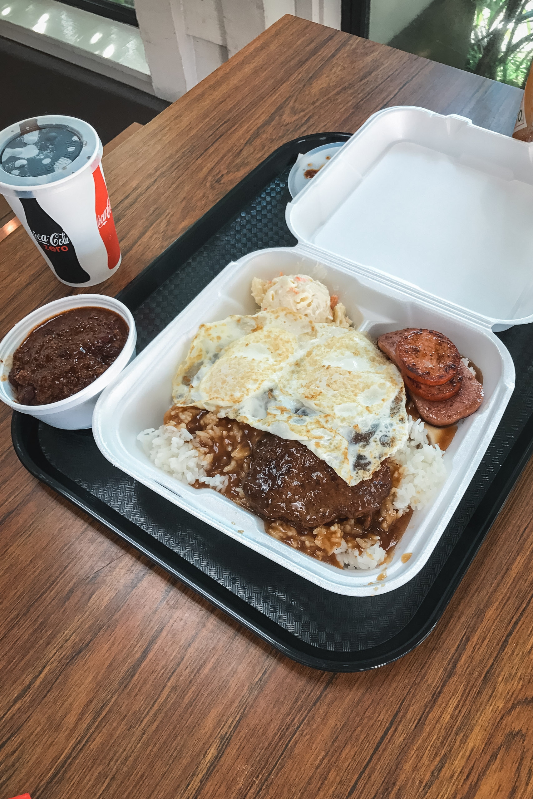 A white takeout container holds a breakfast plate with scrambled eggs, hash browns, gravy-covered meat, and bacon beside a Coca-Cola cup and sauce container on a wooden table