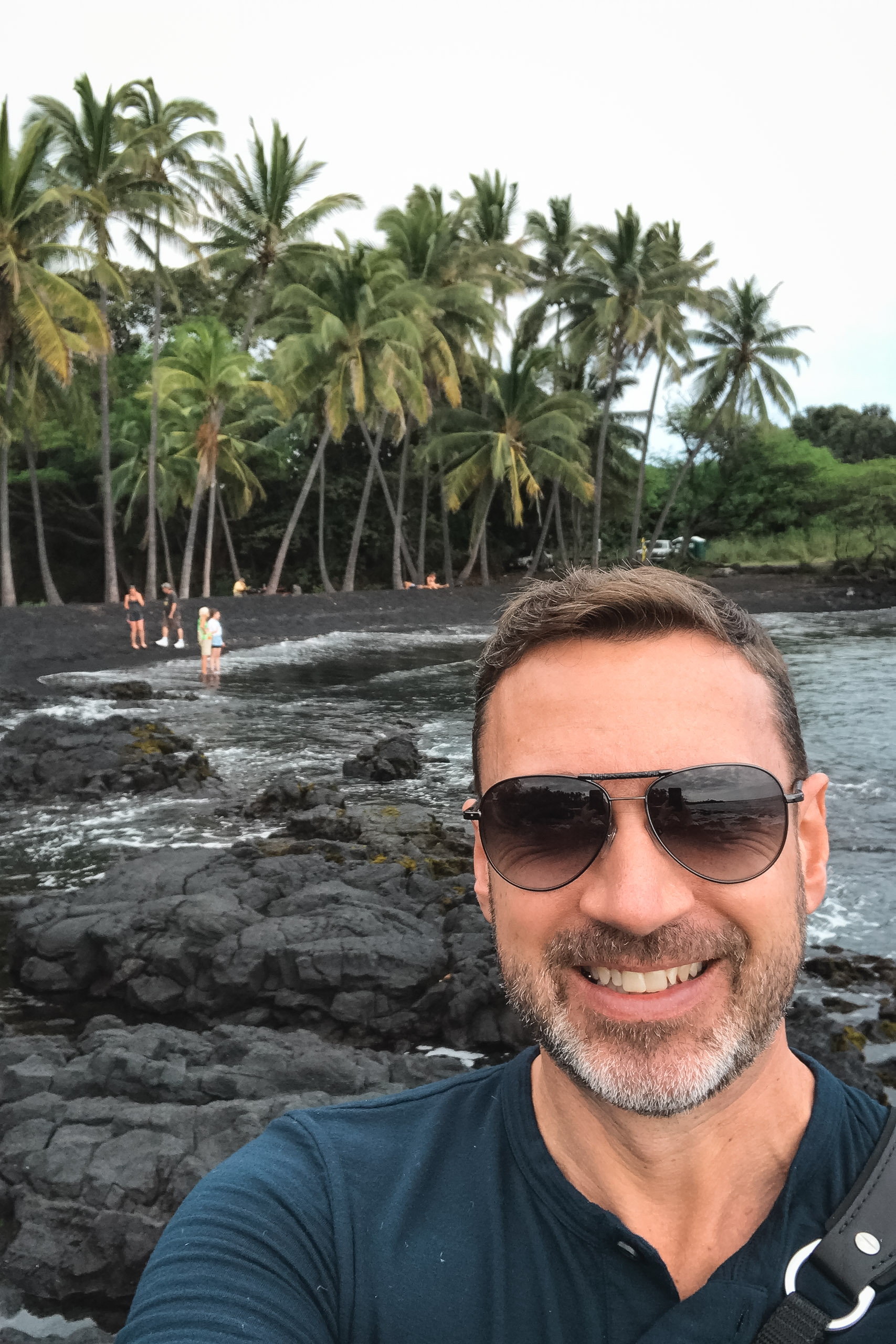 Man wearing sunglasses takes a selfie on a black sand beach with palm trees and wading visitors visible across the water behind him