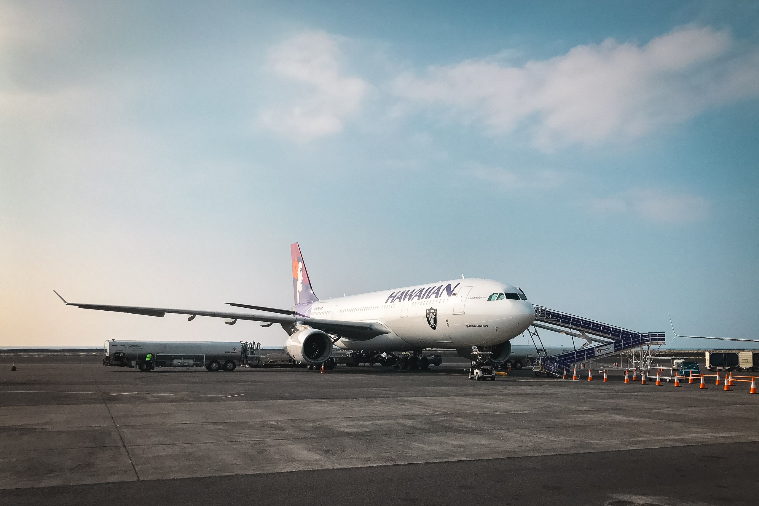 Hawaiian Airlines aircraft parked at gate with ground support vehicles and boarding stairs under clear blue sky with light clouds