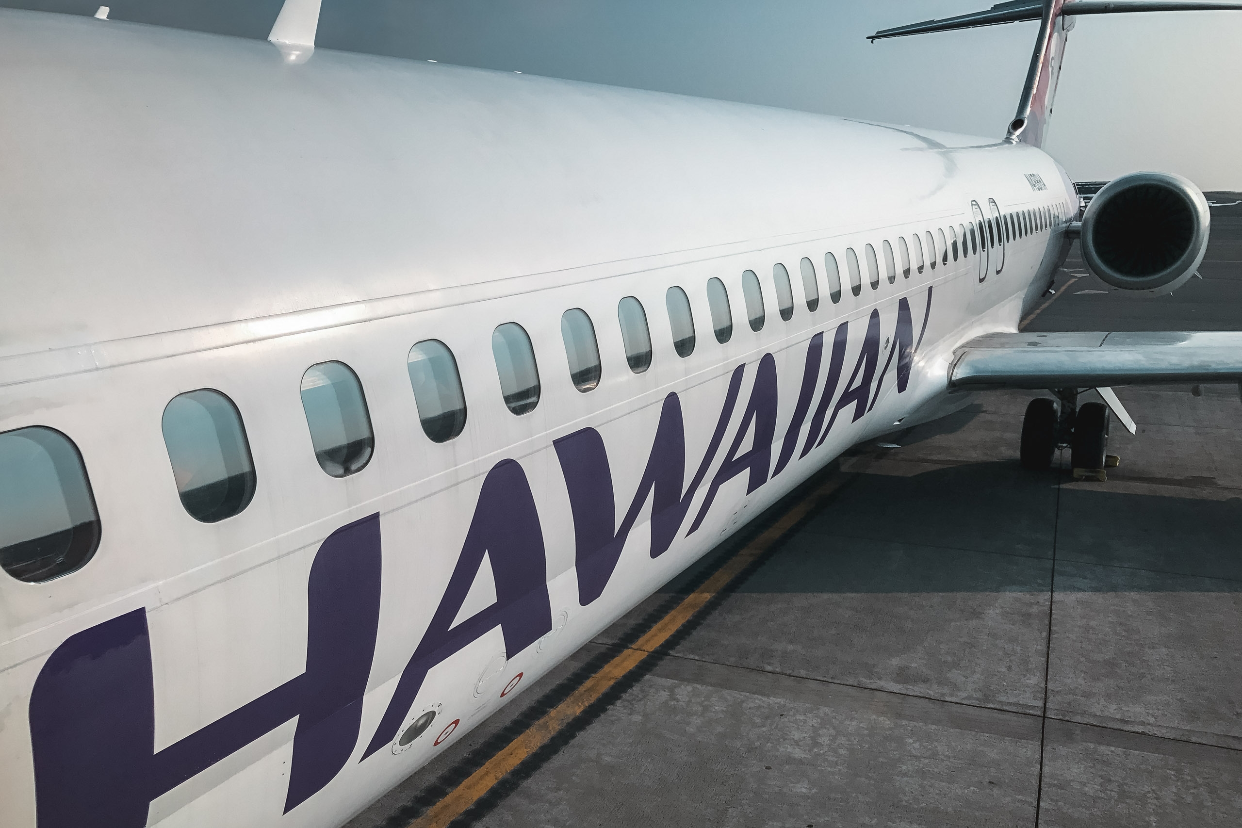 Hawaiian Airlines aircraft sits grounded at the tarmac, displaying rows of passenger windows and navy blue lettering along its white fuselage under bright daylight