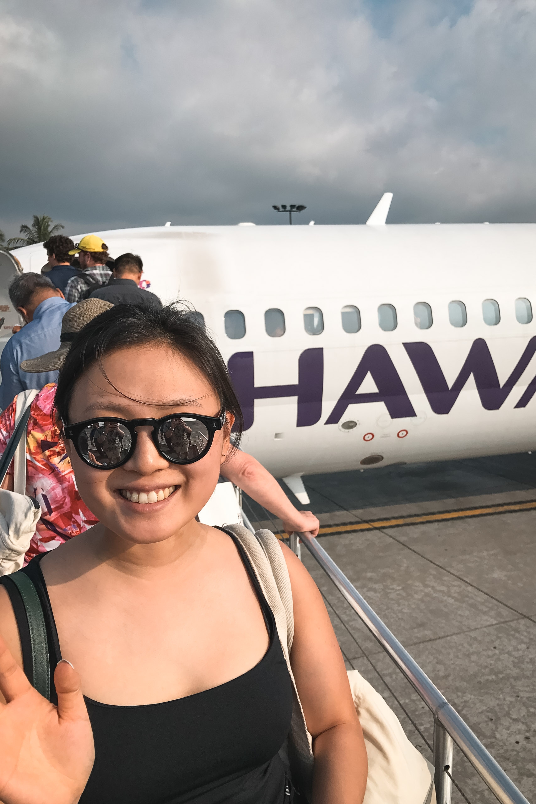 A smiling woman wearing sunglasses stands on airplane stairs boarding a Hawaiian Airlines aircraft under overcast skies at the tarmac