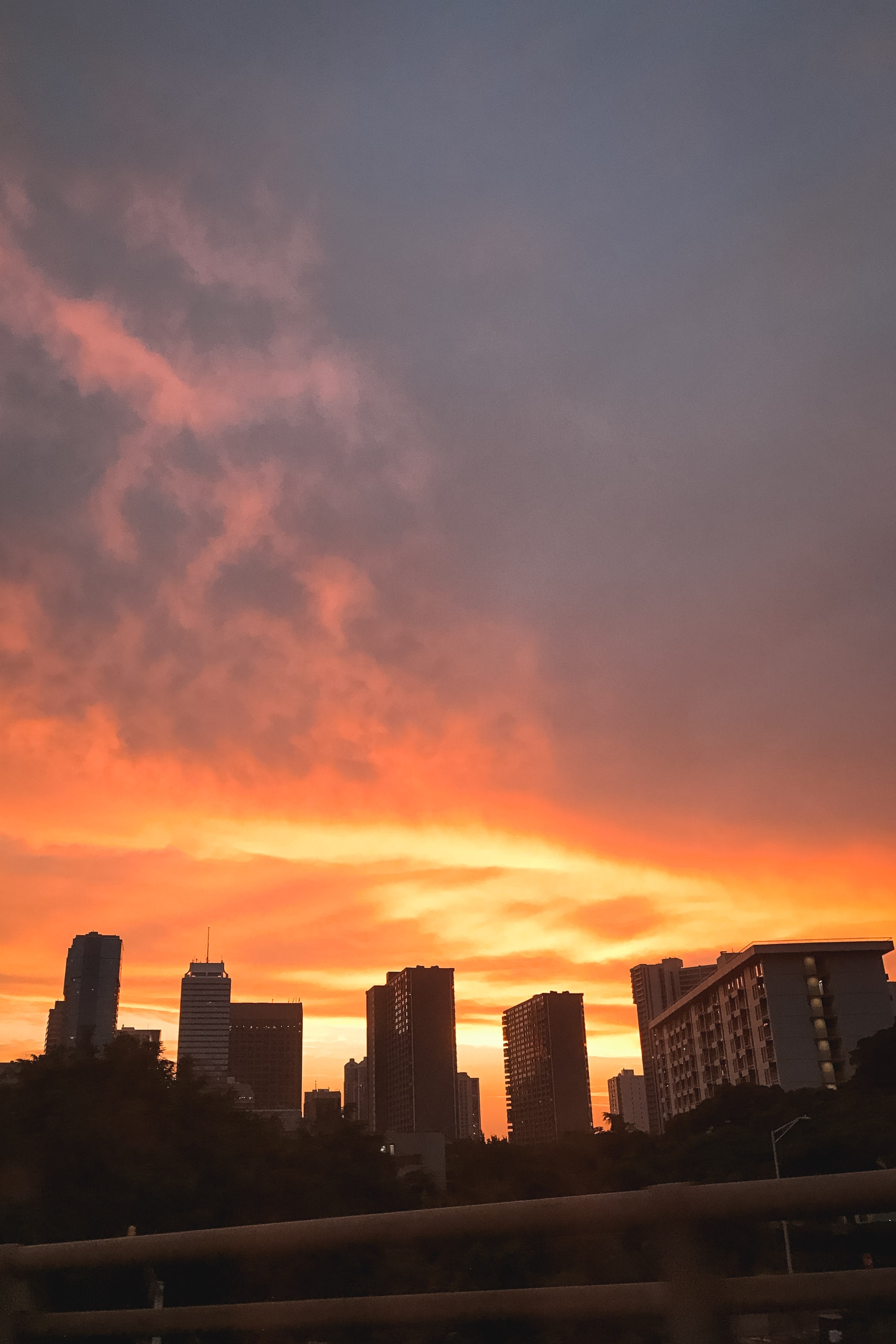 A vibrant orange and pink sunset illuminates dramatic clouds above a silhouetted urban skyline with tall buildings and an overpass in the foreground