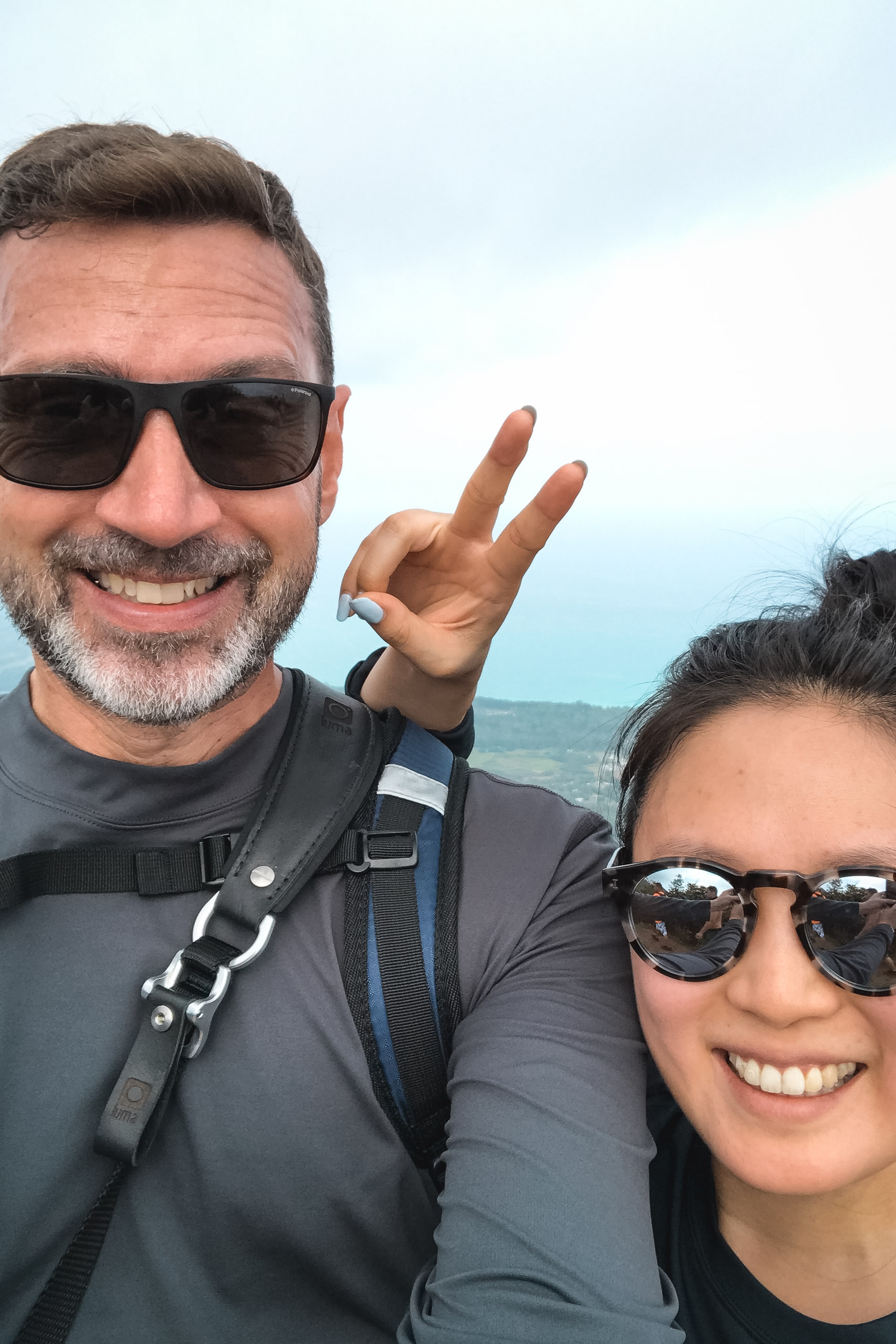 Two people wearing sunglasses and wetsuits smile at the camera against a bright ocean and sky backdrop, with the man making a peace sign