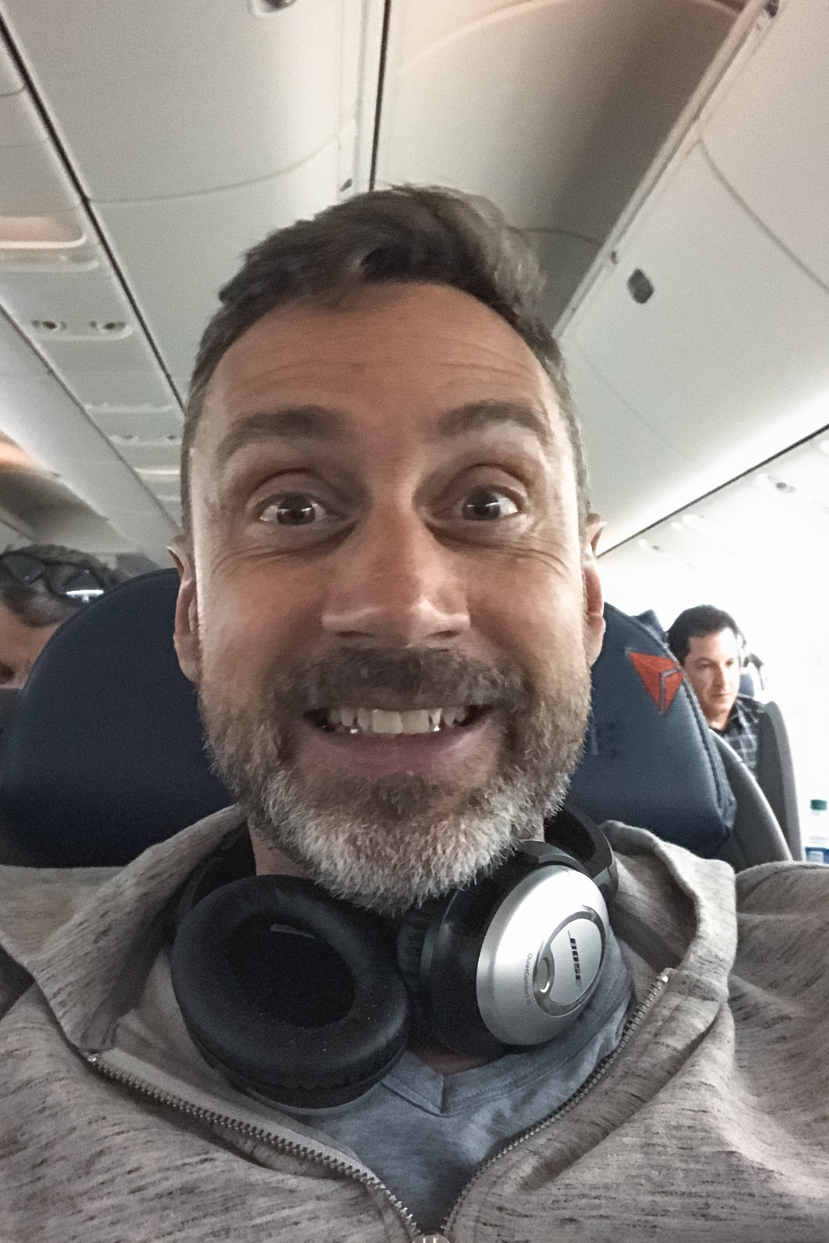 Bearded man wearing headphones on his chest smiles widely at camera inside an airplane cabin with passengers visible in background