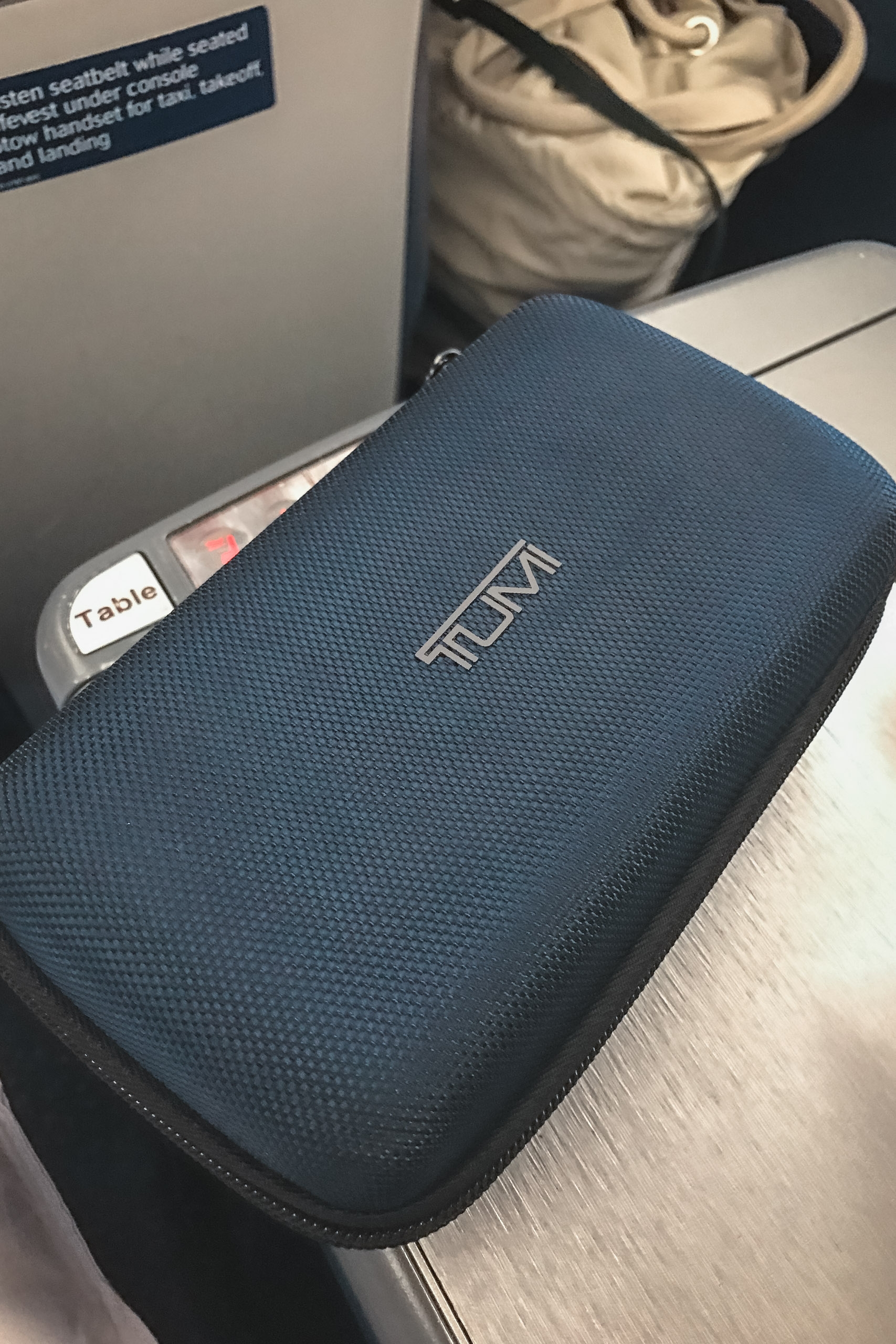 A blue TUMI travel case sits on an airplane seat with a "Table" label visible nearby, captured in overhead cabin lighting during flight
