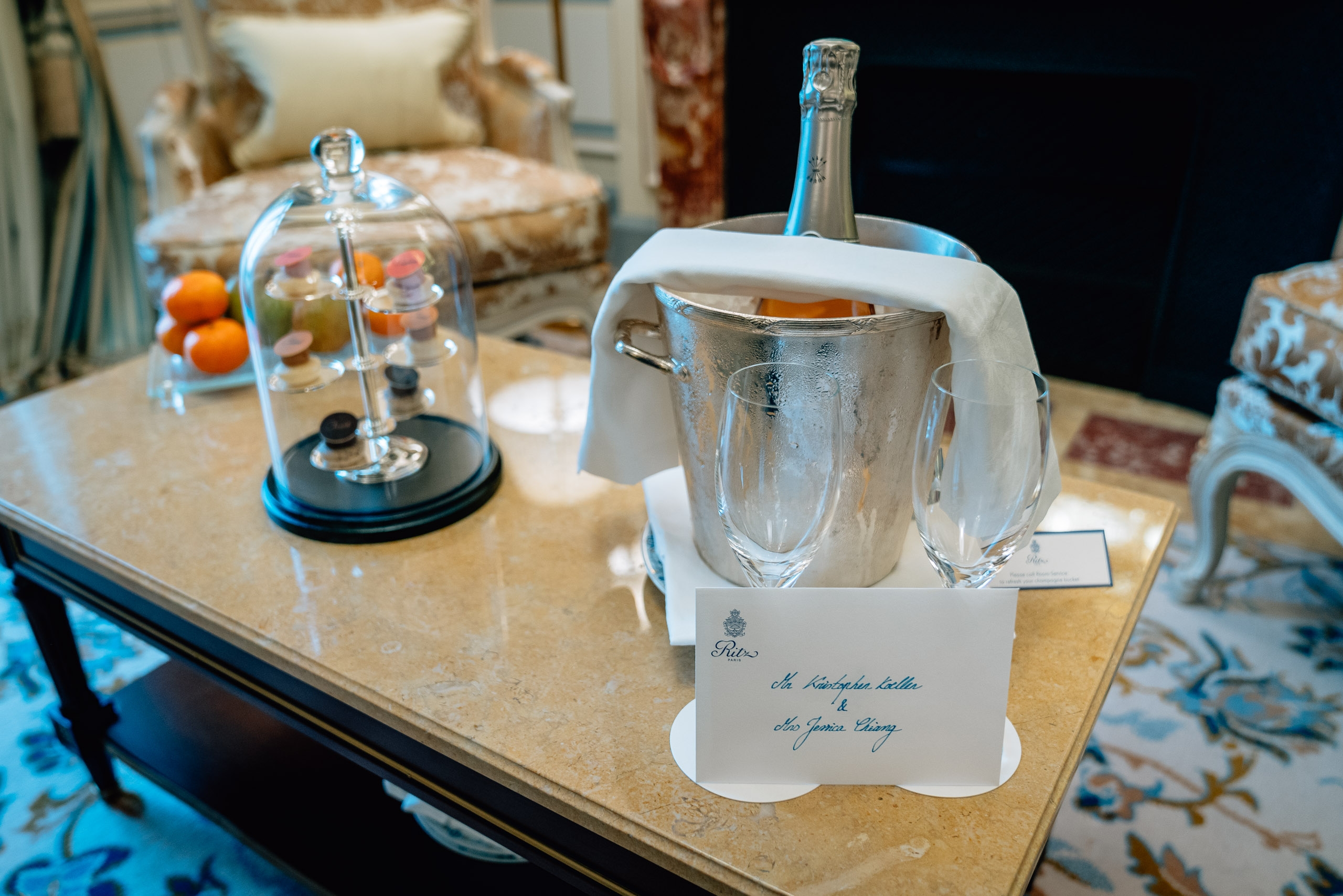 Luxurious hotel suite detail featuring a marble table set with champagne bucket, crystal glasses, decorative cloche with colorful macarons, and formal place card in an elegantly appointed parlor room