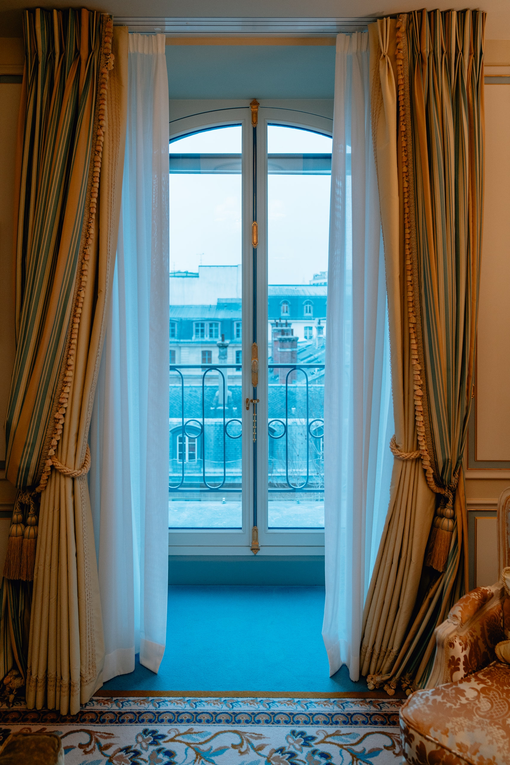 French doors with striped gold and cream curtains frame a Parisian balcony overlooking classical blue-roofed buildings and ornate ironwork railings in soft daylight