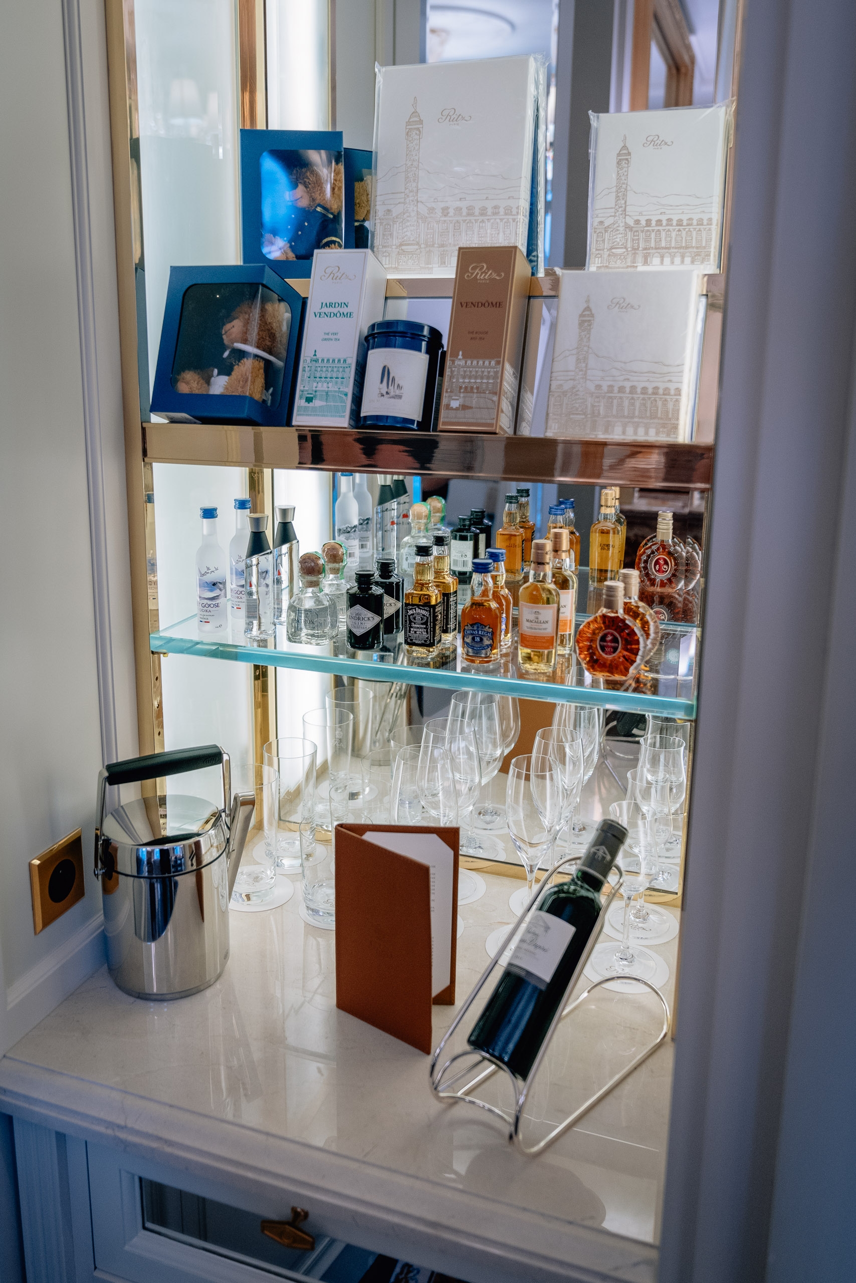 A glass display cabinet showcases luxury fragrances and spirits with architectural illustrations of Parisian buildings, organized across multiple shelves with elegant glassware and decorative items below