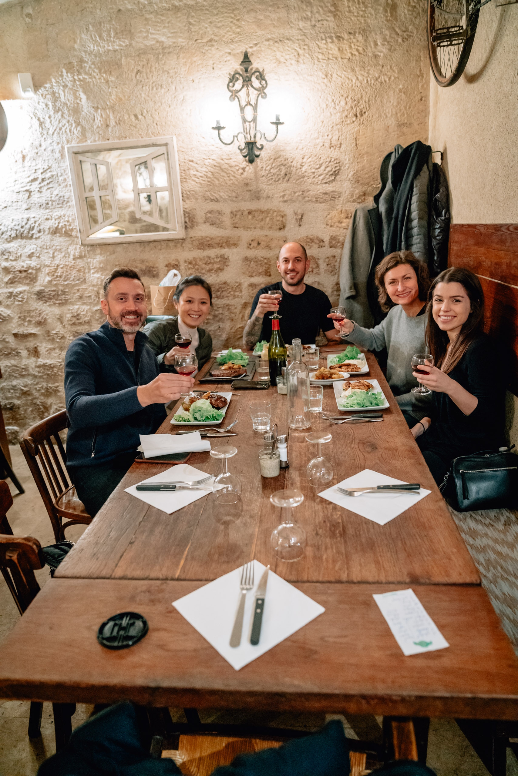 Five people gather around a wooden dining table in a rustic stone-walled room, raising glasses and smiling while enjoying a meal together under warm candlelit ambiance