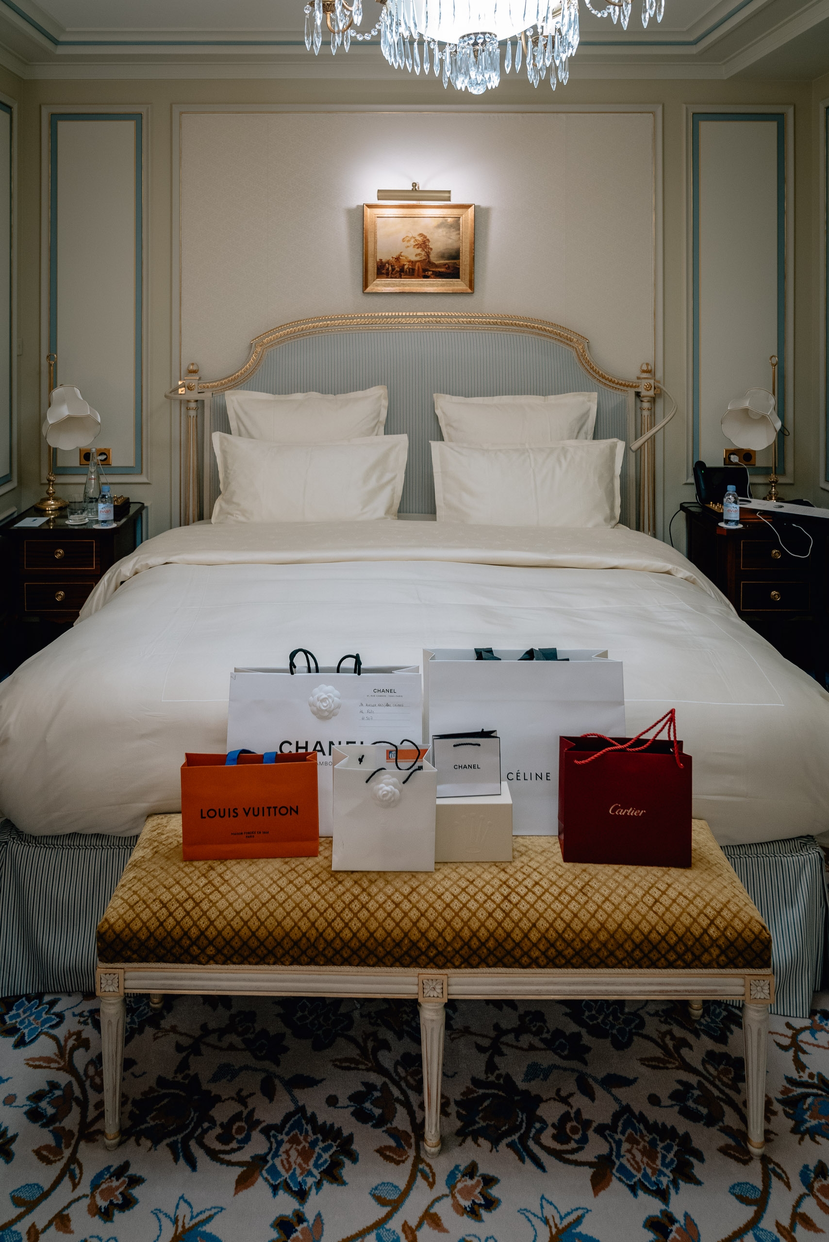 Elegant bedroom featuring a gilded bed with cream linens, luxury shopping bags arranged on a patterned bench, crystal chandelier overhead, and symmetrical dark nightstands beneath framed artwork