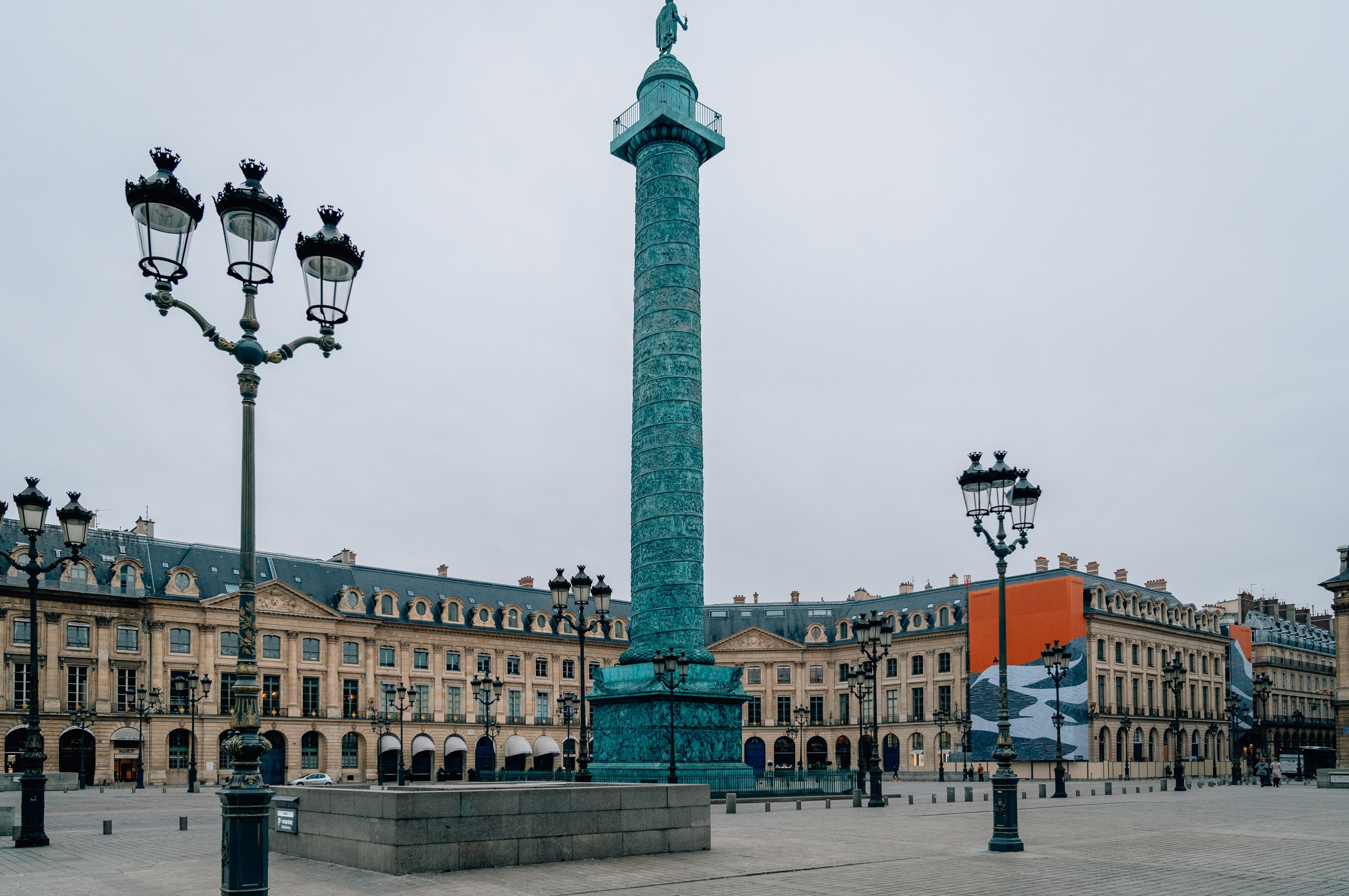 A grand Parisian plaza features an ornate green column monument surrounded by elegant classical buildings with mansard roofs and ornamental street lamps under overcast skies