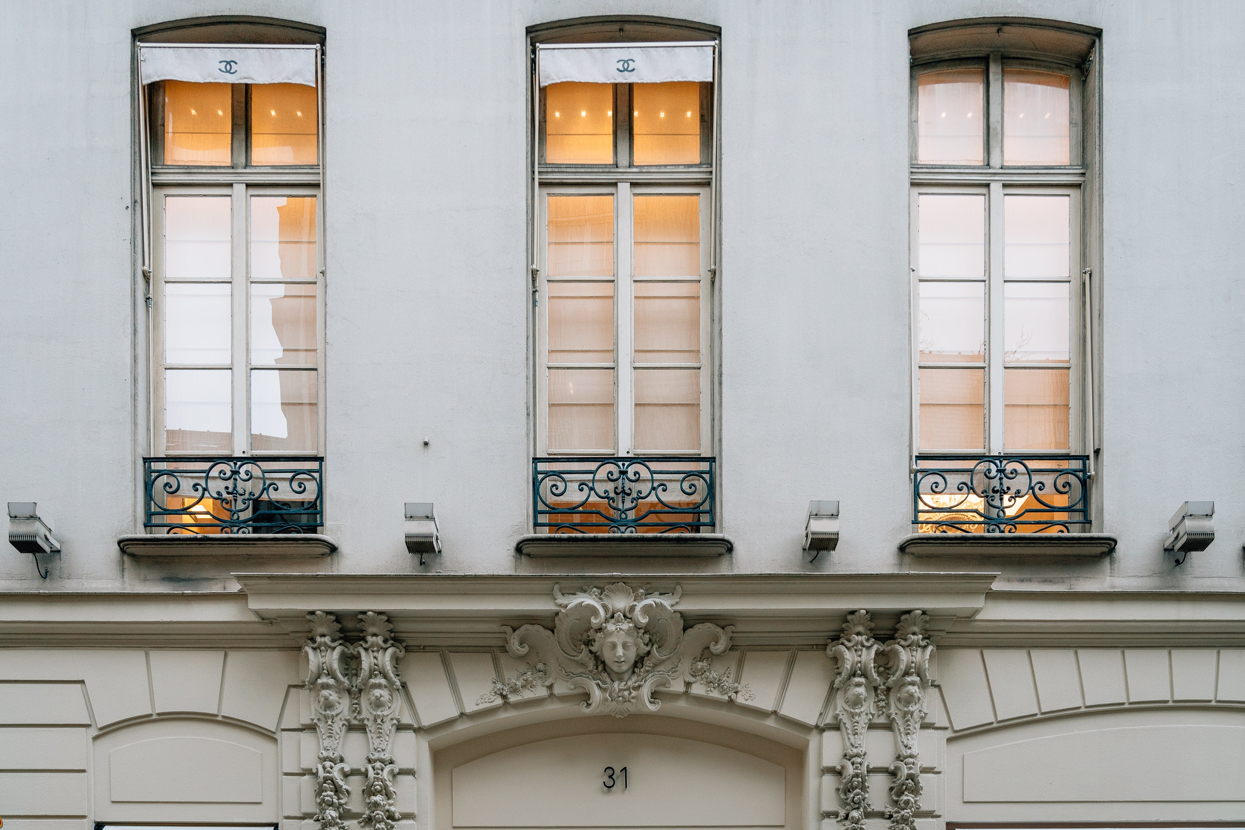 Three tall Parisian apartment windows with ornate iron balconies glow warmly against a pale stone facade, crowned by classical sculptural details and the number 31