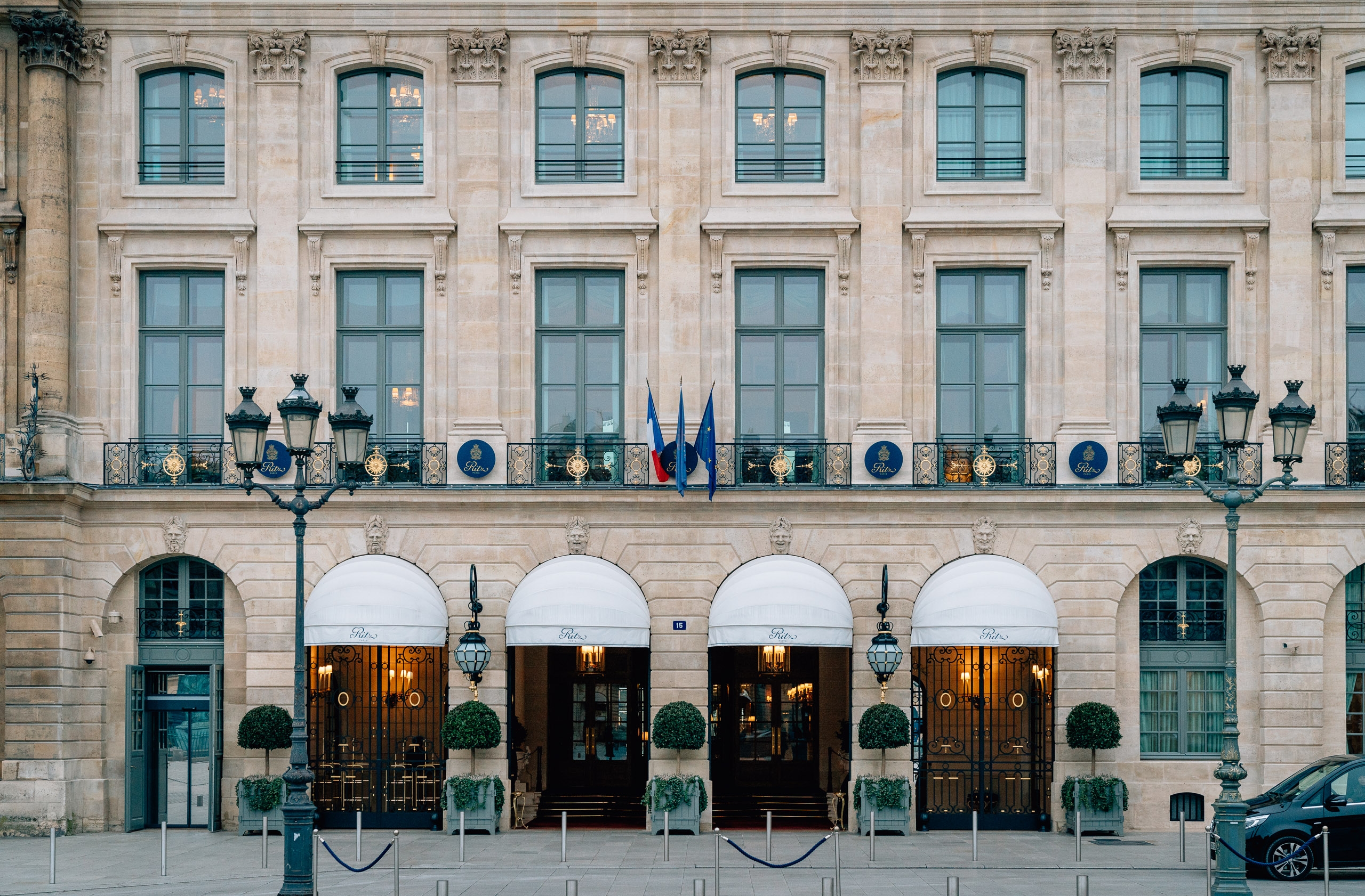 Classical Parisian facade of the Ritz hotel features symmetrical arched entrances with white awnings, ornate street lamps, and rows of tall windows beneath decorative cornices