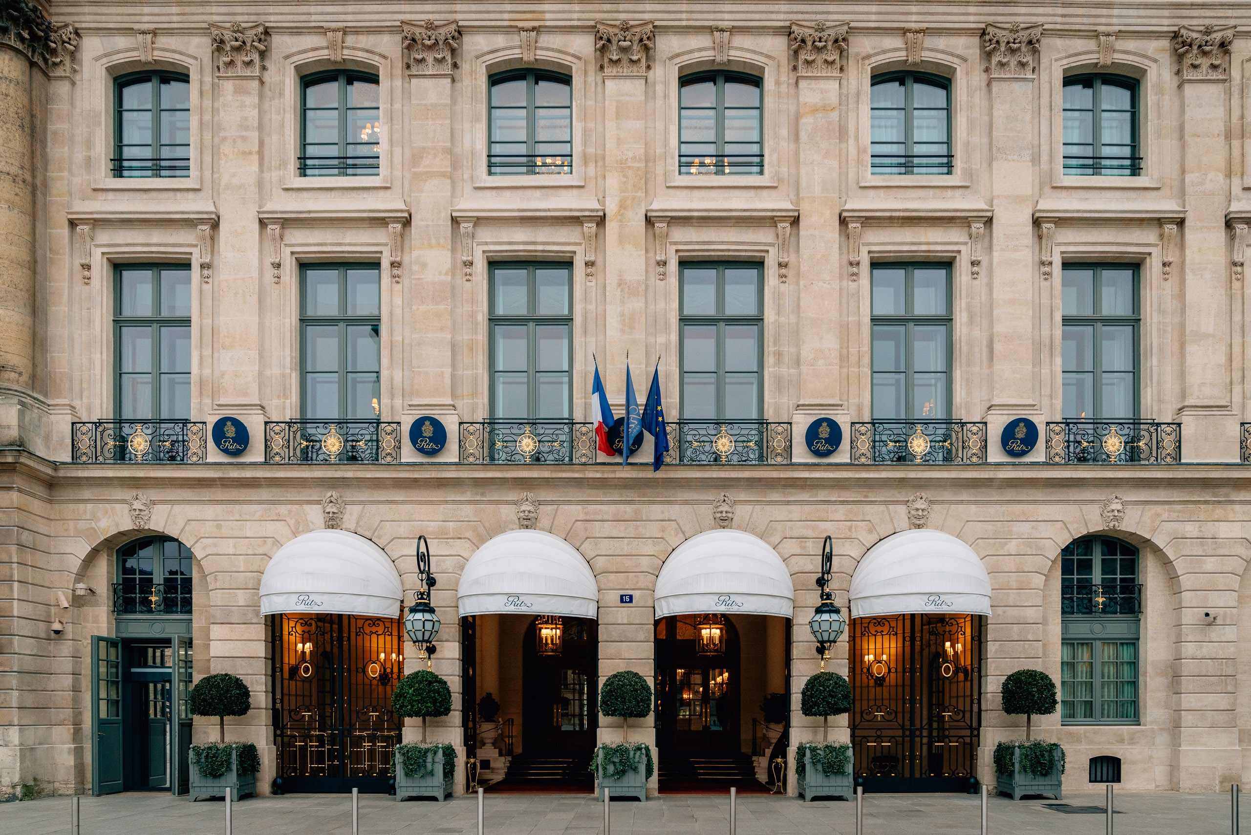 The Ritz Paris hotel facade displays symmetrical Beaux-Arts architecture with cream stone, rows of tall windows, ornate capitals, and a grand ground-floor entrance featuring white awnings and manicured topiaries beneath French flags