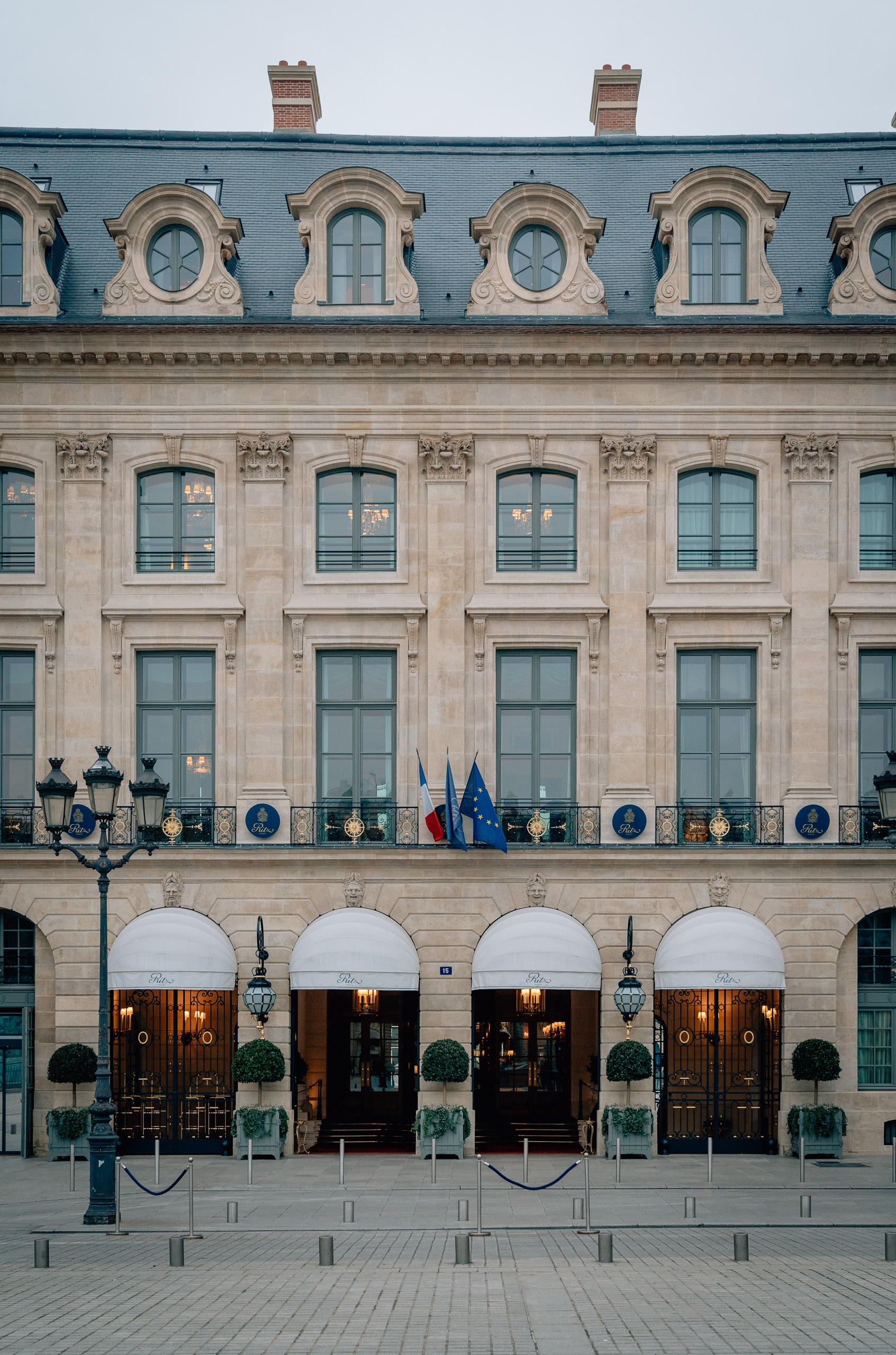 The Ritz Paris facade displays classical Parisian architecture with cream-colored stone, mansard roof, and symmetrical arched entrances flanked by ornate lampposts and potted topiaries